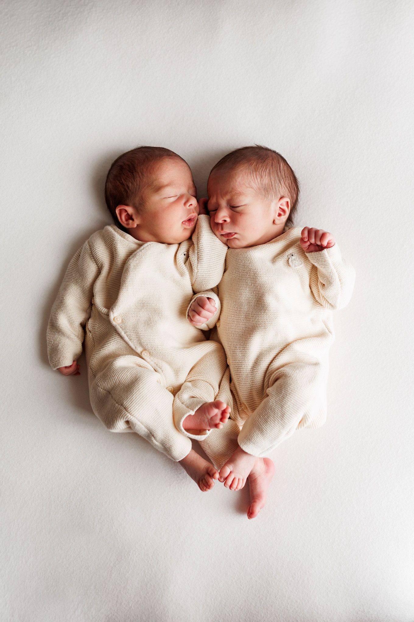 Two sleeping newborn twins in cream knit outfits lie cuddled side by side on a soft white blanket, heads touching.