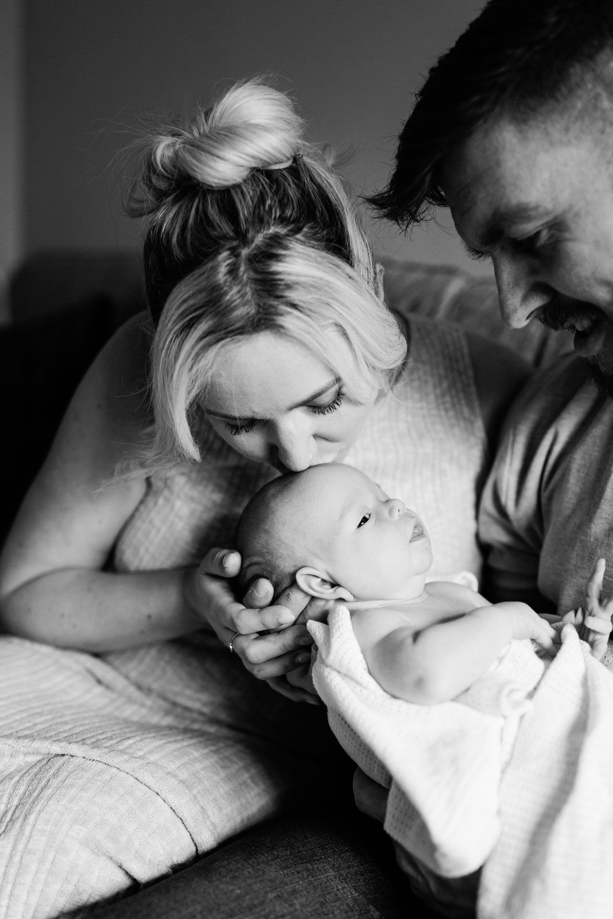 Black and white photo of parents lovingly gazing at their newborn baby, mother kissing infant's head.