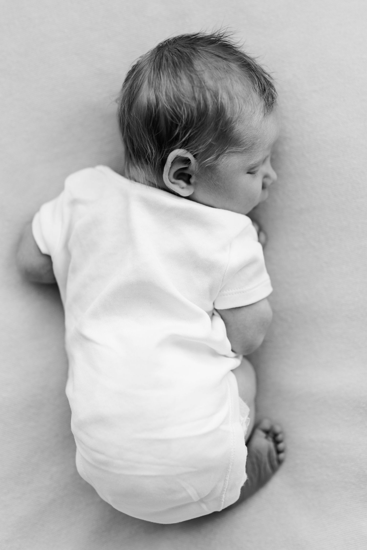 Black and white photo of a sleeping newborn baby in a white onesie lying on their side.