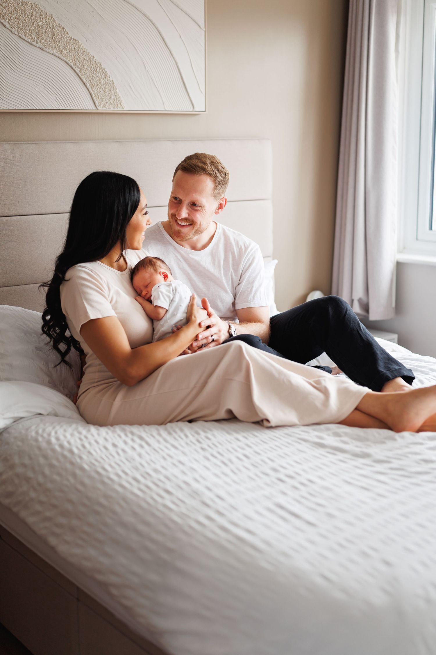A smiling couple sits together on a white bed in a bright bedroom, gently cradling their sleeping newborn baby between them.