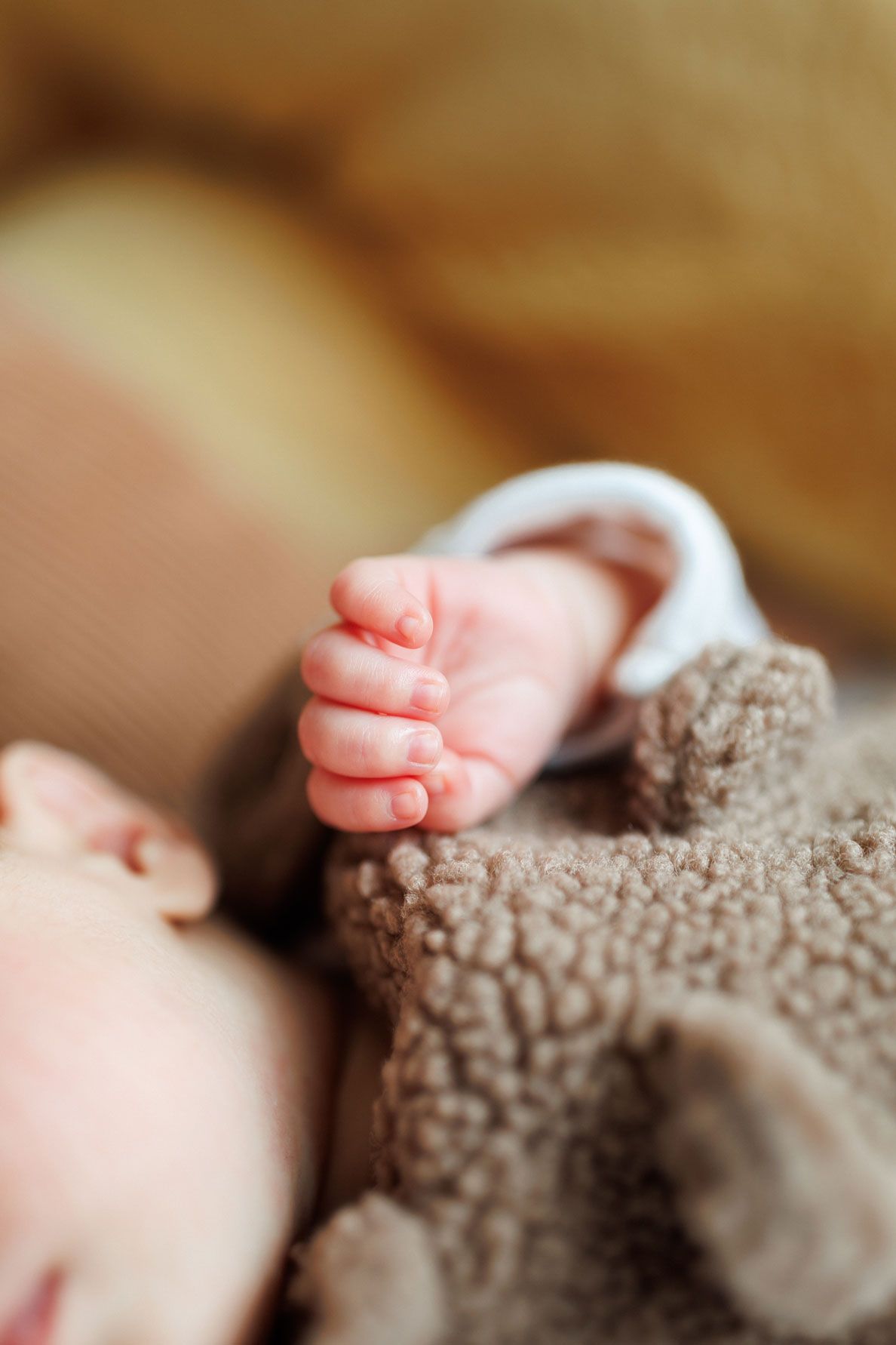 Close-up of a newborn baby's tiny curled hand resting on a soft brown teddy bear fleece outfit, with the sleeping baby softly blurred.