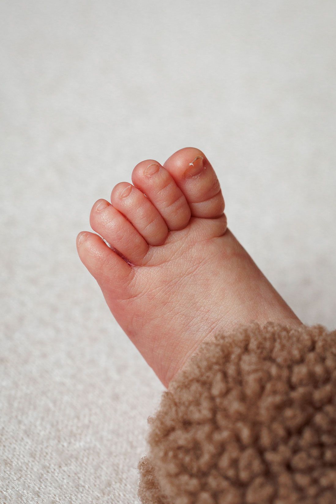 Close-up of a newborn baby's tiny foot with curled toes beside a soft brown teddy fleece sleeve on a cream blanket.