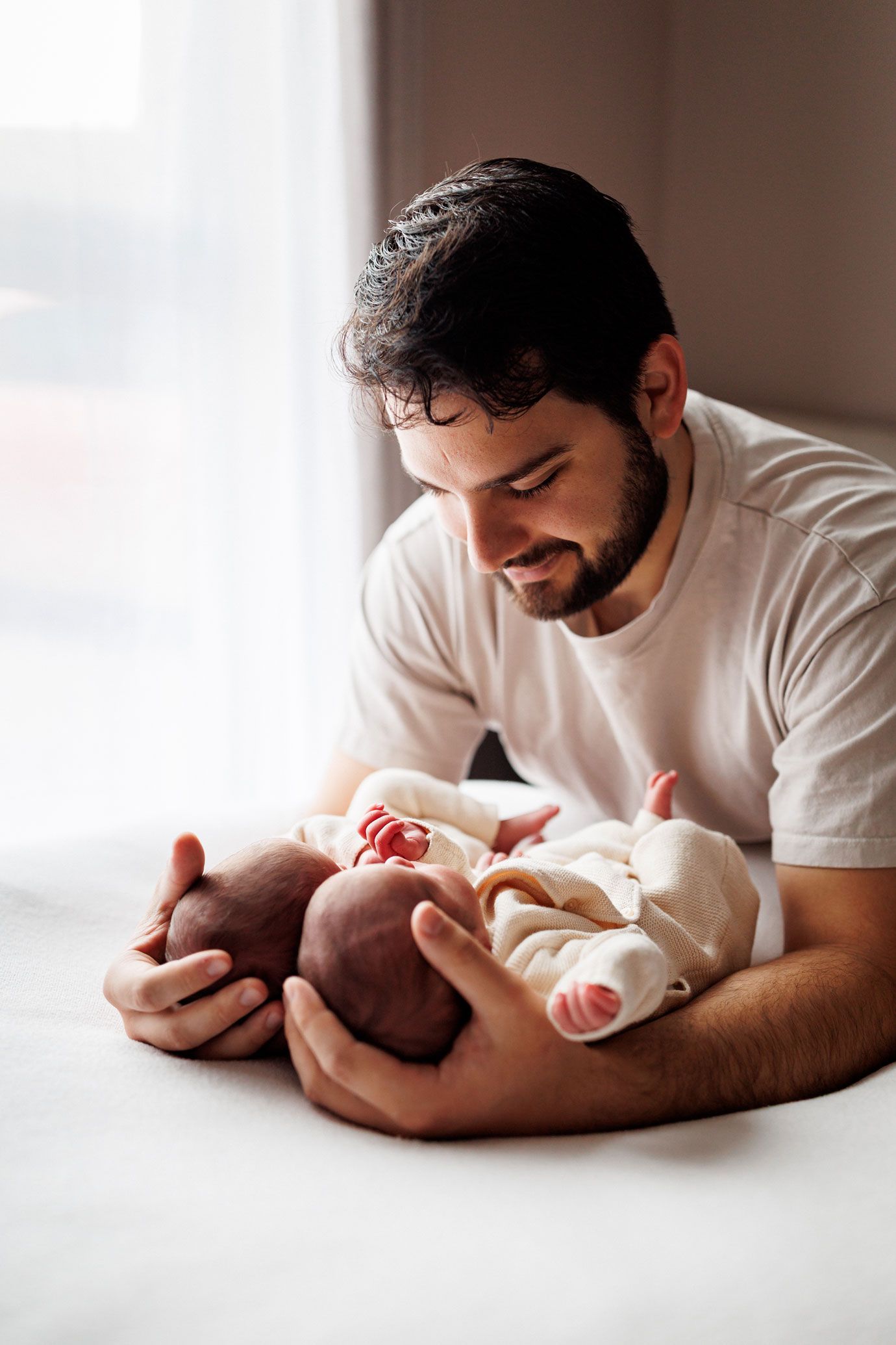 A smiling bearded father in a white t-shirt gazes lovingly at his two swaddled newborn twins resting on a soft bed.