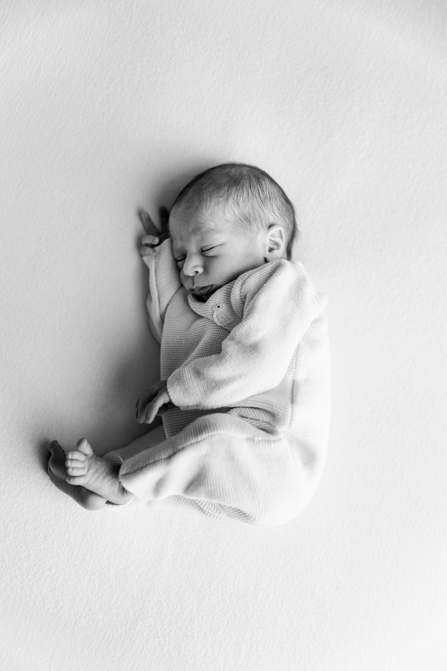 Black and white photo of a sleeping newborn baby in a soft knit outfit, curled up peacefully on a smooth light blanket.