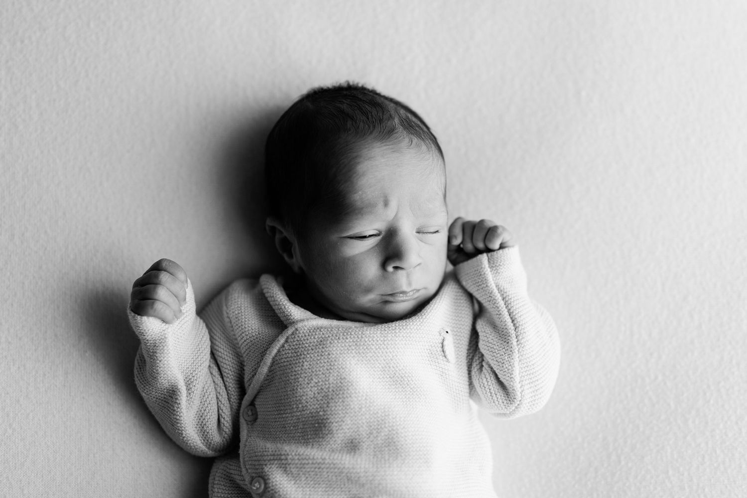 Black and white portrait of a sleeping newborn baby in a soft knit outfit with tiny fists raised beside the face.