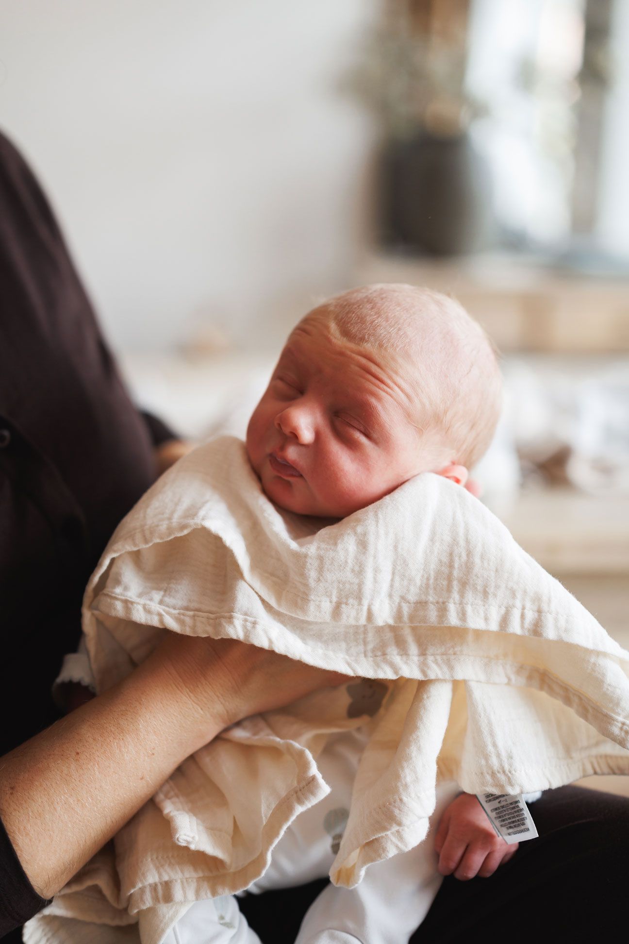Sleeping newborn baby wrapped in a cream muslin swaddle blanket, cradled in an adult's arms in a softly lit, cozy room.
