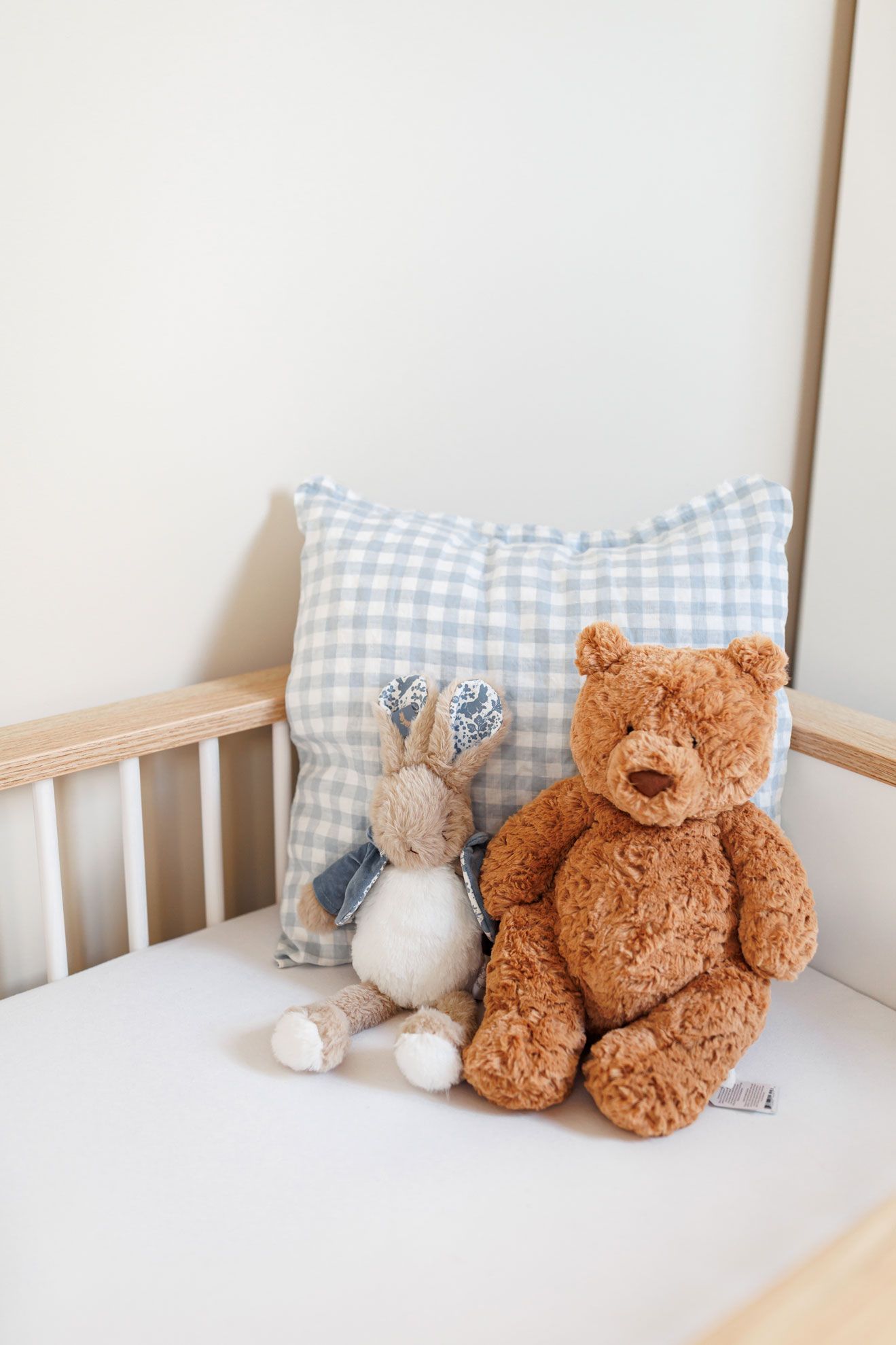 A brown teddy bear and a plush bunny sit beside a blue gingham cushion in a wooden baby cot.
