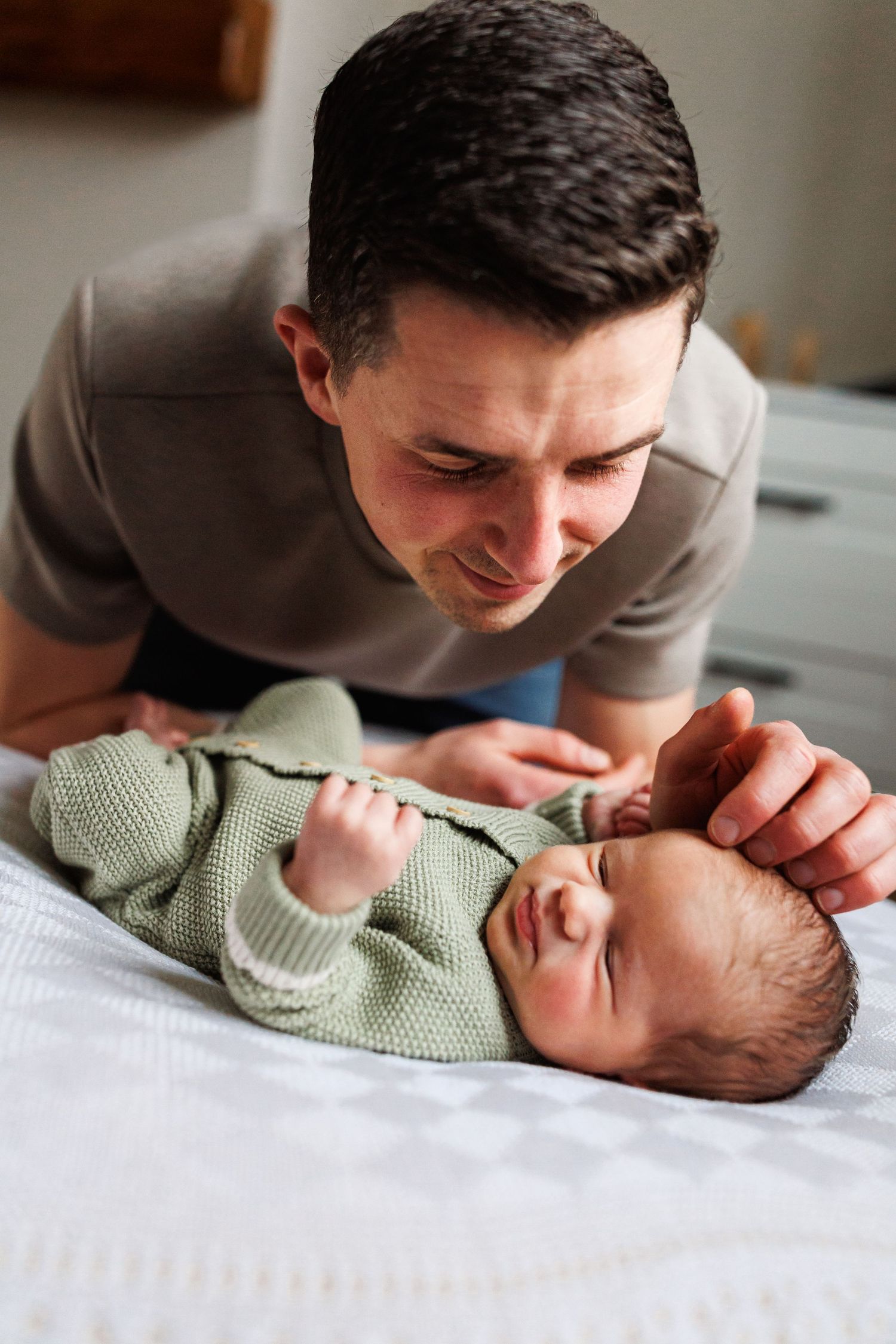 A young father in a beige shirt smiles tenderly at his newborn baby in a green knit outfit lying on a bed.