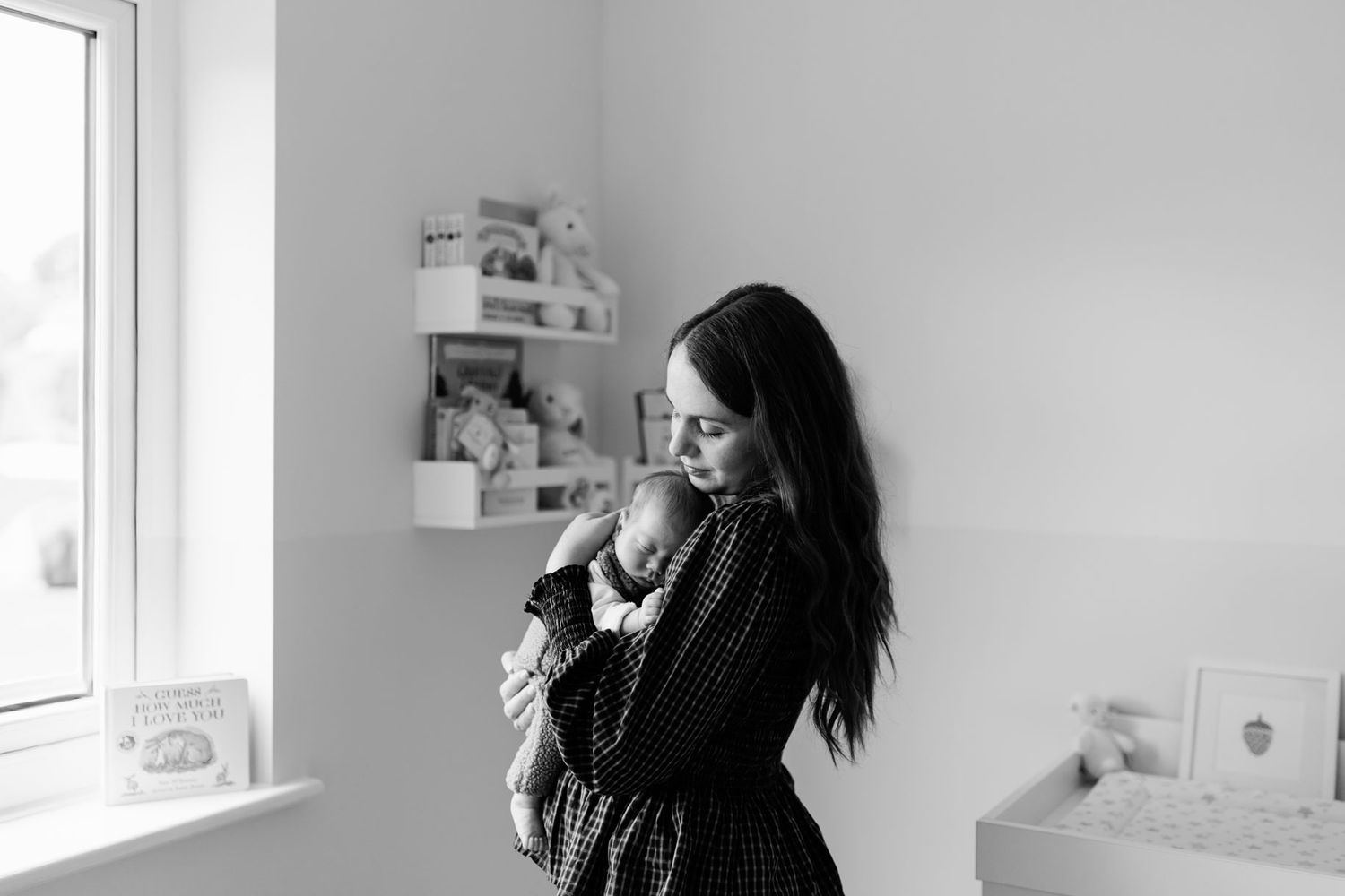Black and white photo of a mother with long dark hair cradling a sleeping newborn baby beside a sunlit window in a cozy nursery.
