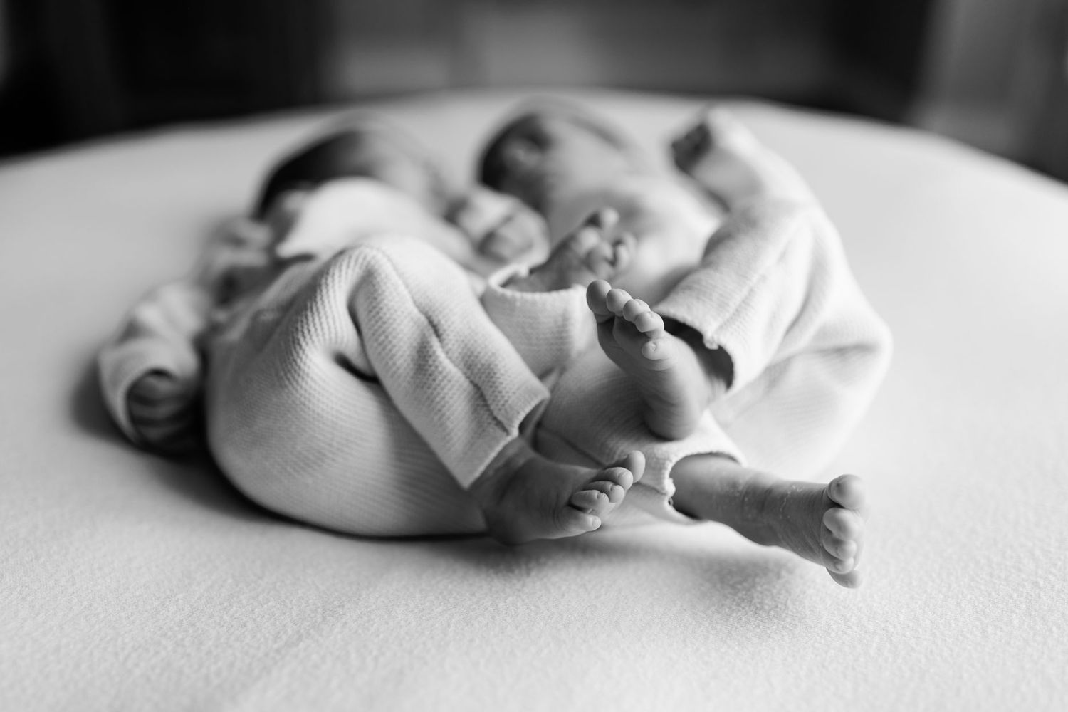 Black and white photo of two sleeping newborn twins in soft knit outfits cuddled together, with tiny bare feet in sharp focus.