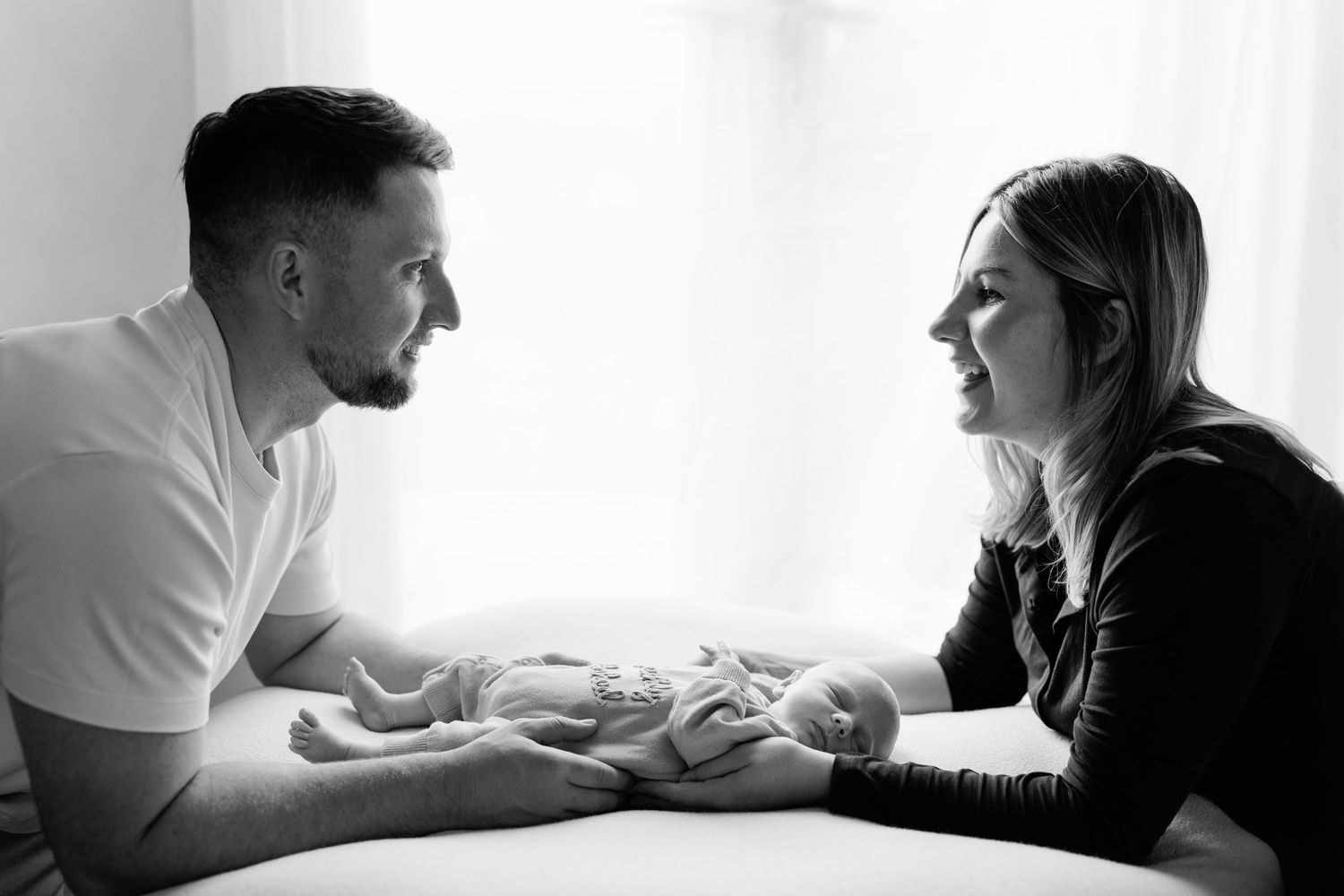 Black and white newborn photo of a smiling man and woman facing each other with their hands gently cradling a sleeping baby.