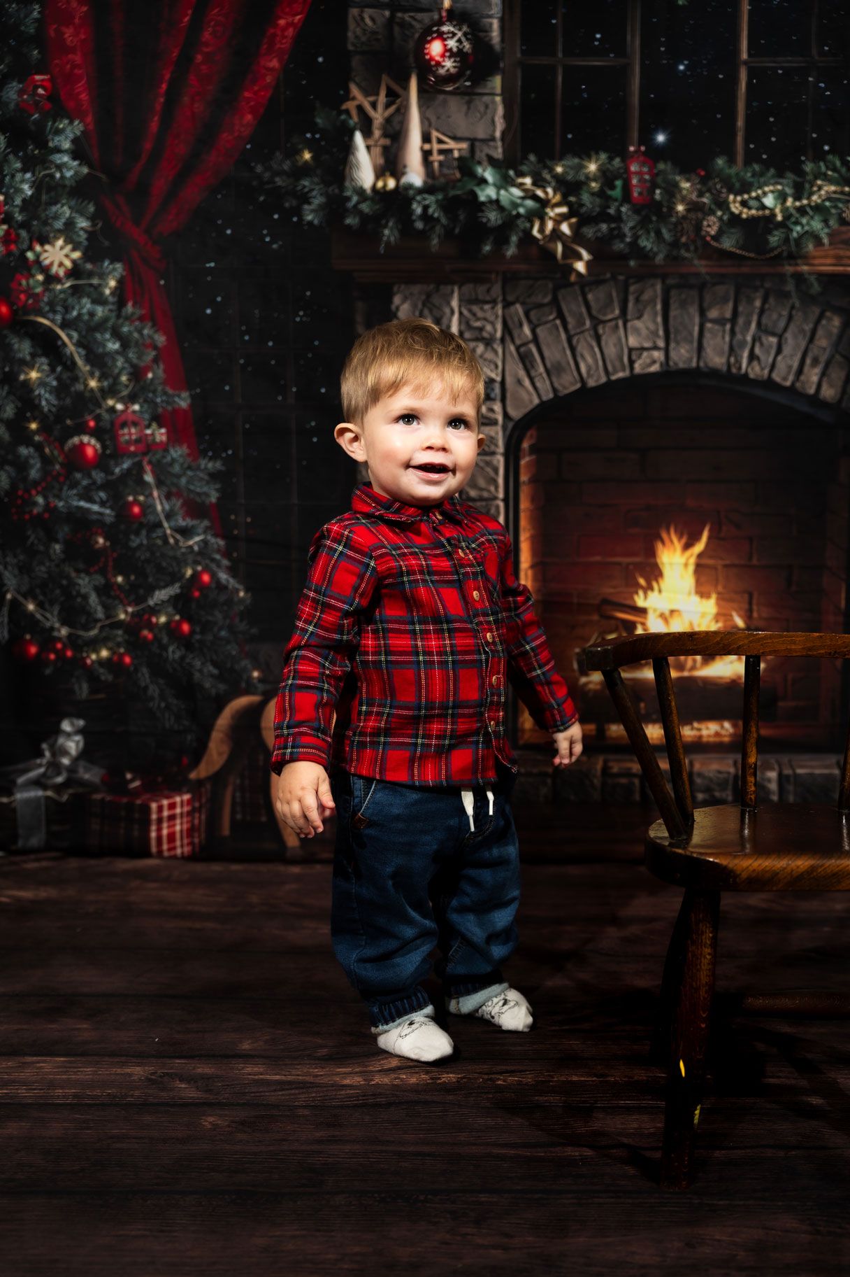 Toddler in a red plaid shirt smiling beside a glowing fireplace and decorated Christmas tree in a cozy holiday portrait.