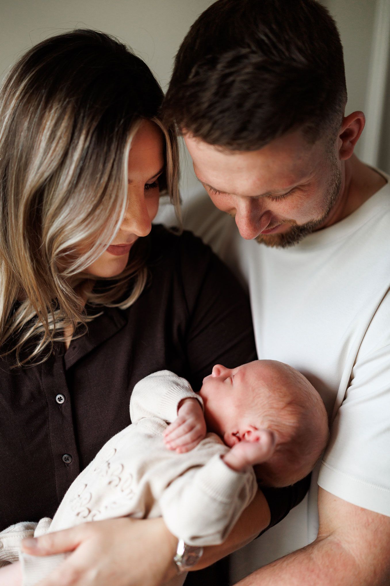 A smiling mother and father gaze lovingly down at their sleeping newborn baby, wrapped in a cream knit outfit, in a warm family moment.