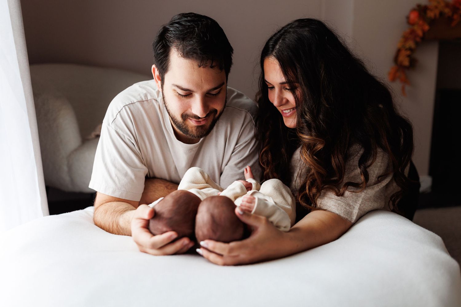 Smiling couple lying on a white bed gazing lovingly at their newborn baby in a cozy, softly lit newborn family photo.