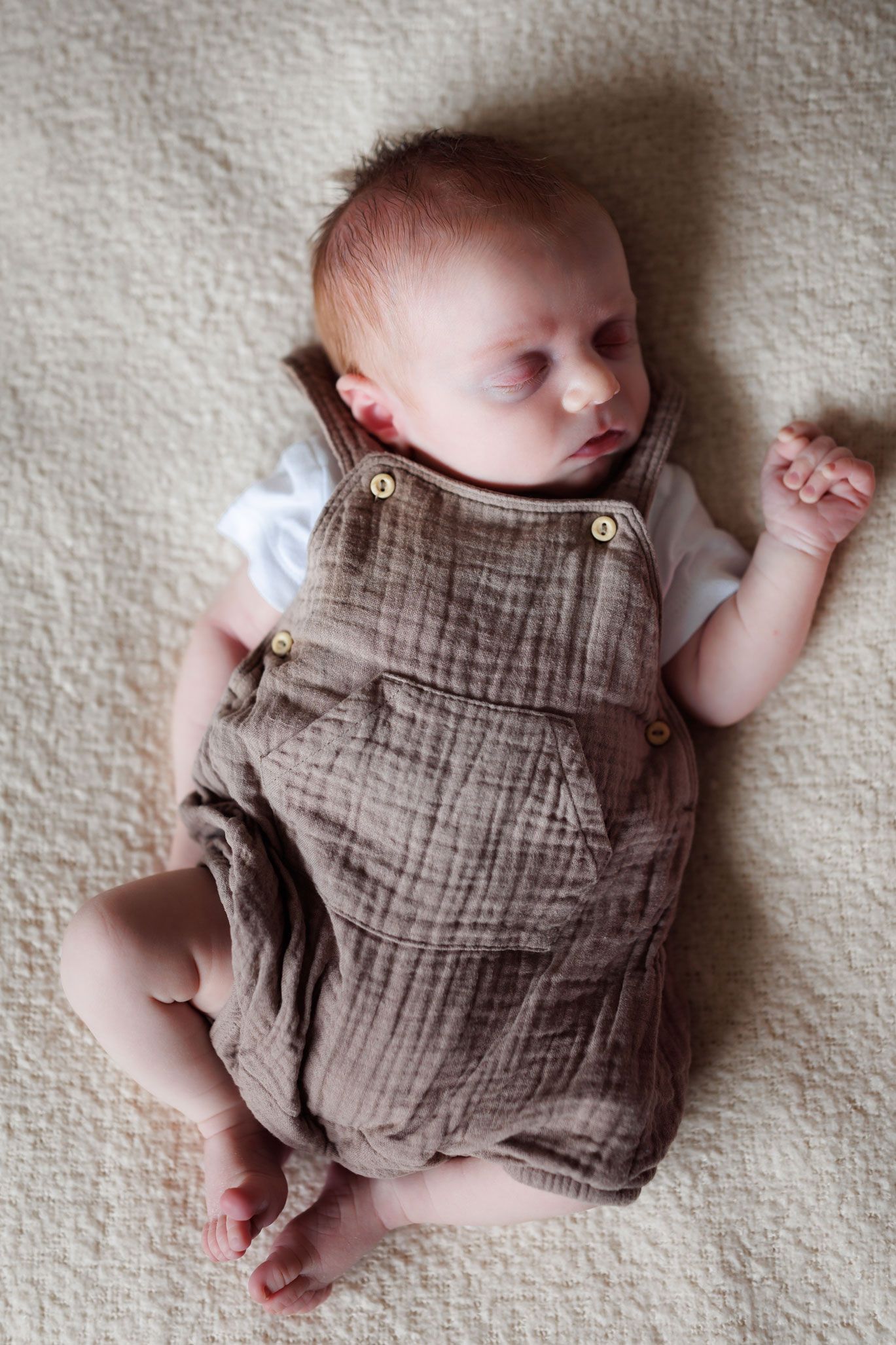 Newborn baby sleeping peacefully on a cream blanket, wearing brown linen overalls with button straps in their Nottingham Newborn Session