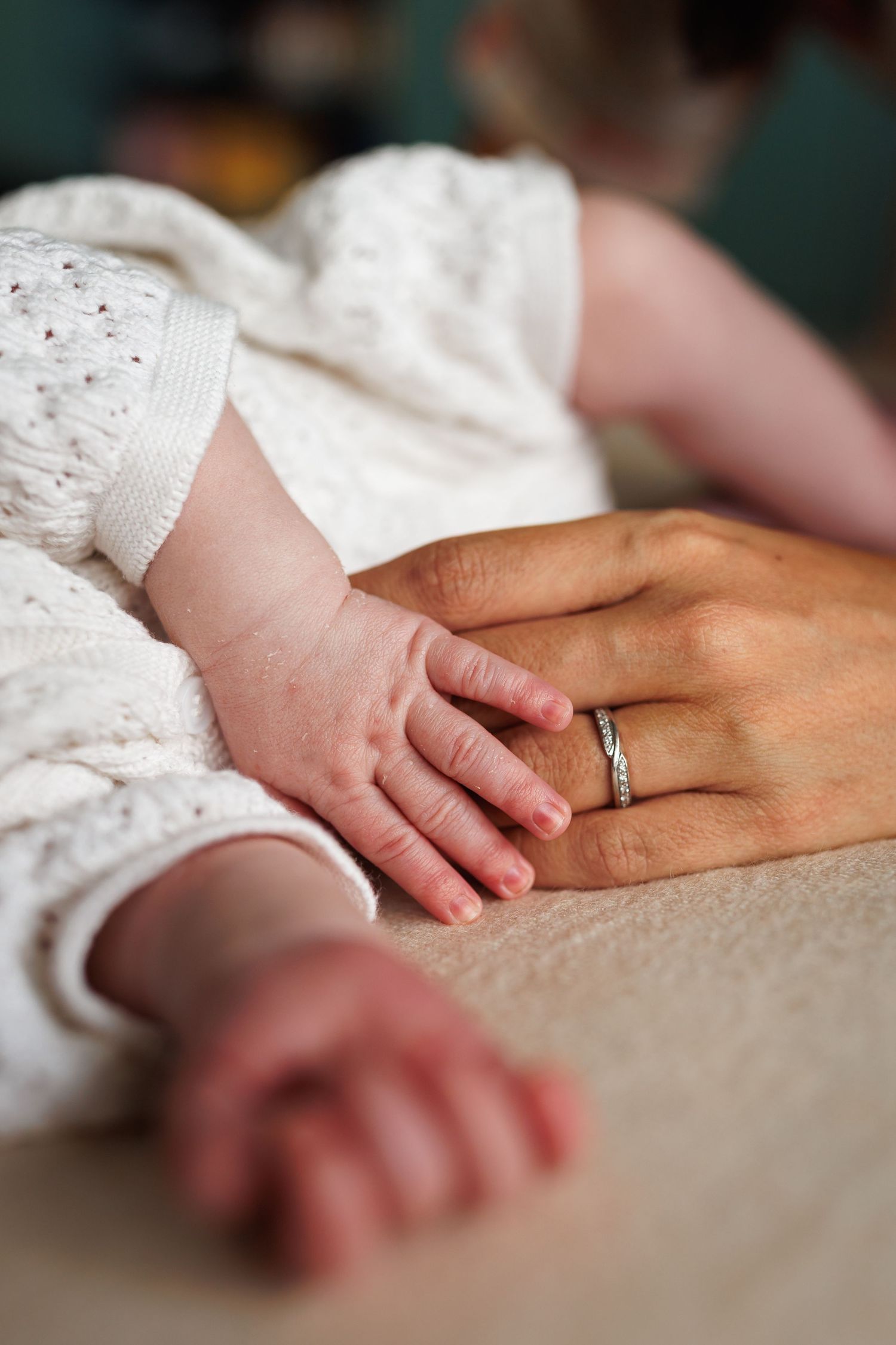 Newborn baby tiny hand resting on mothers ringed hand in white knit sleepsuit.