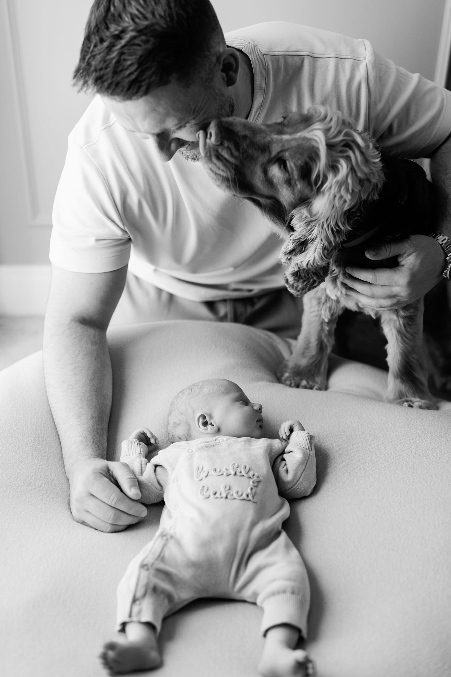 Black and white photo of a smiling father and his fluffy dog leaning over a sleeping newborn baby in a onesie lying on a soft bed.