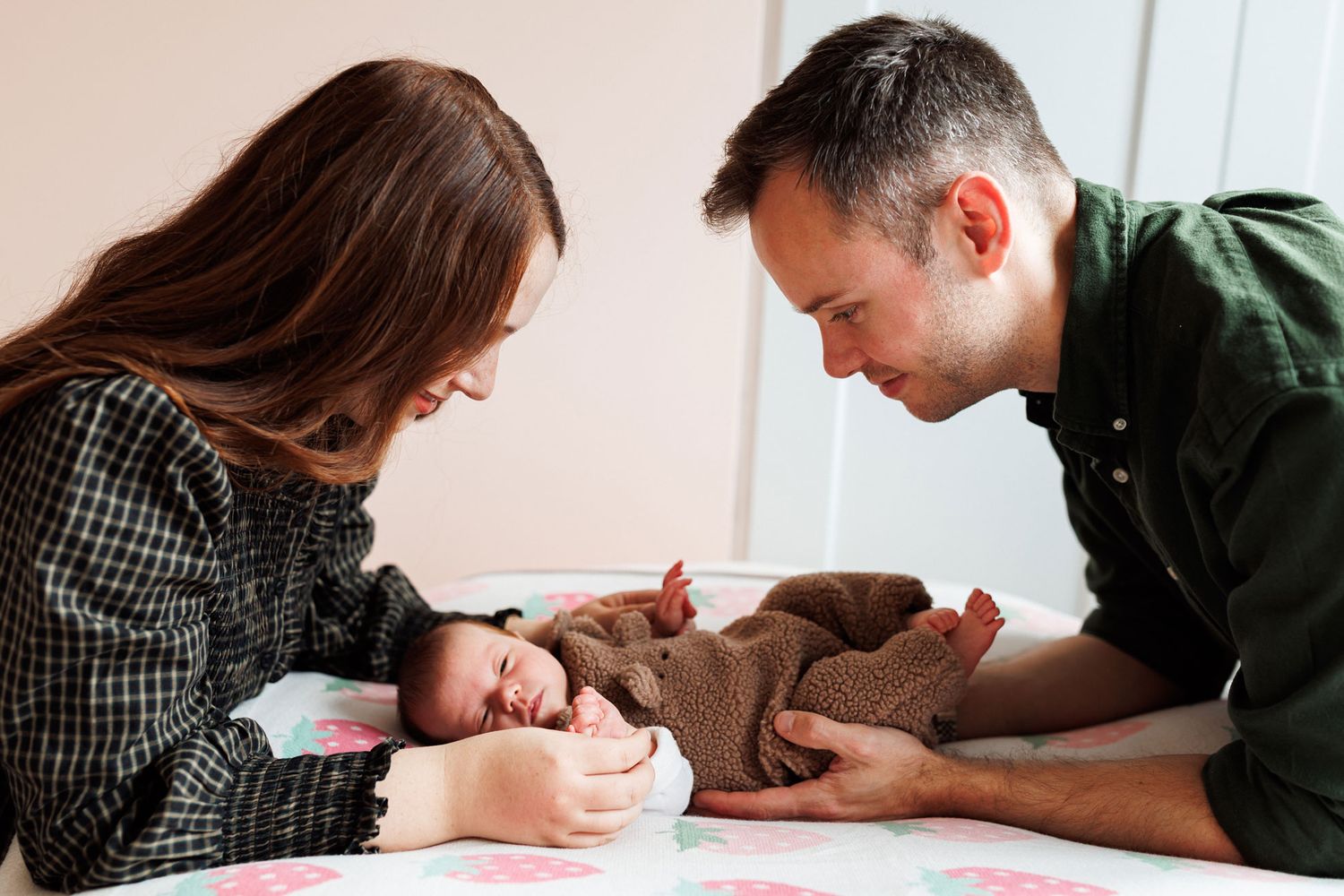 Smiling parents gently touch their sleeping newborn baby dressed in a brown knit outfit, lying on a soft pink floral blanket.