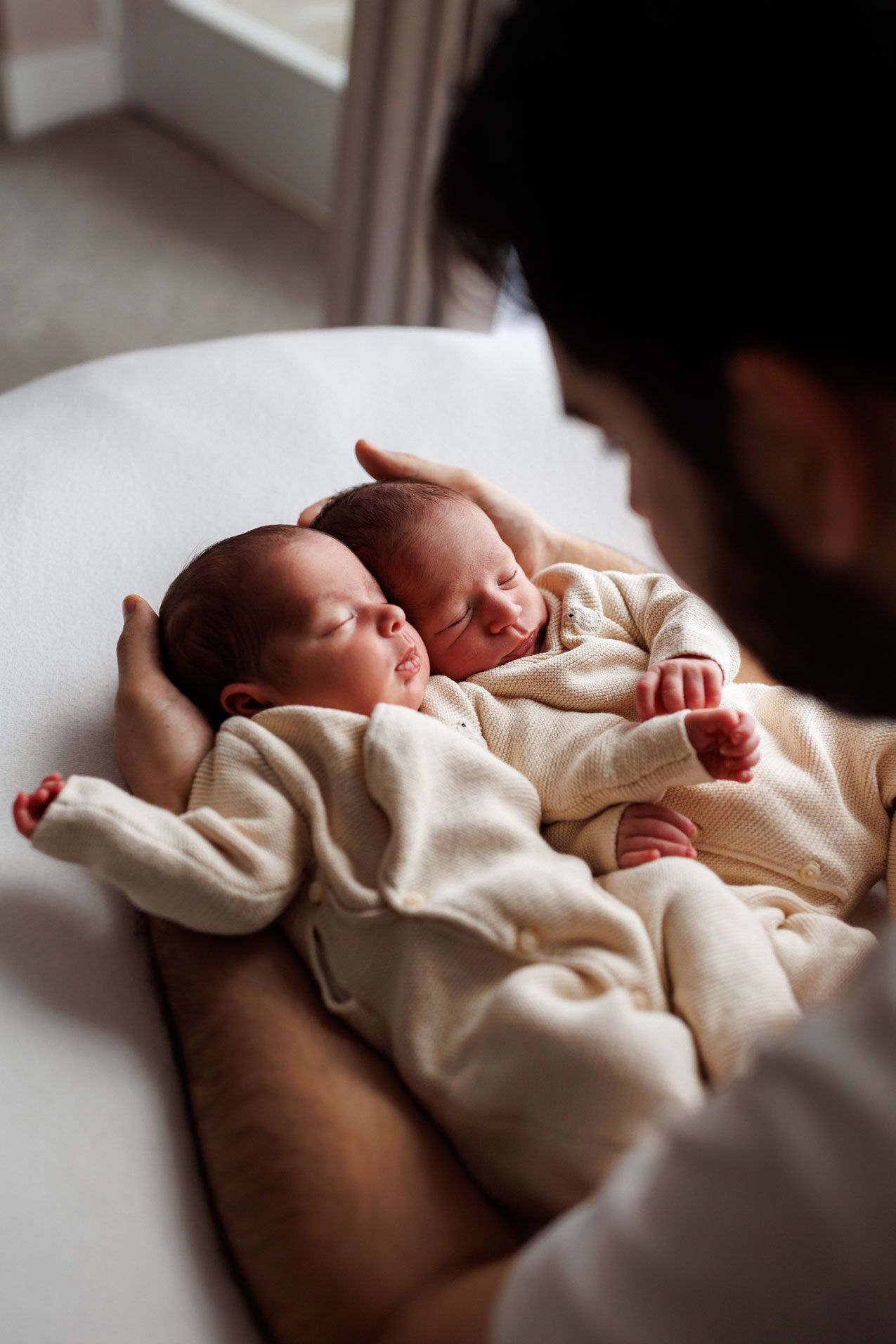 Two sleeping newborn twins in matching cream outfits rest together on a white blanket while a bearded father gently cradles them.