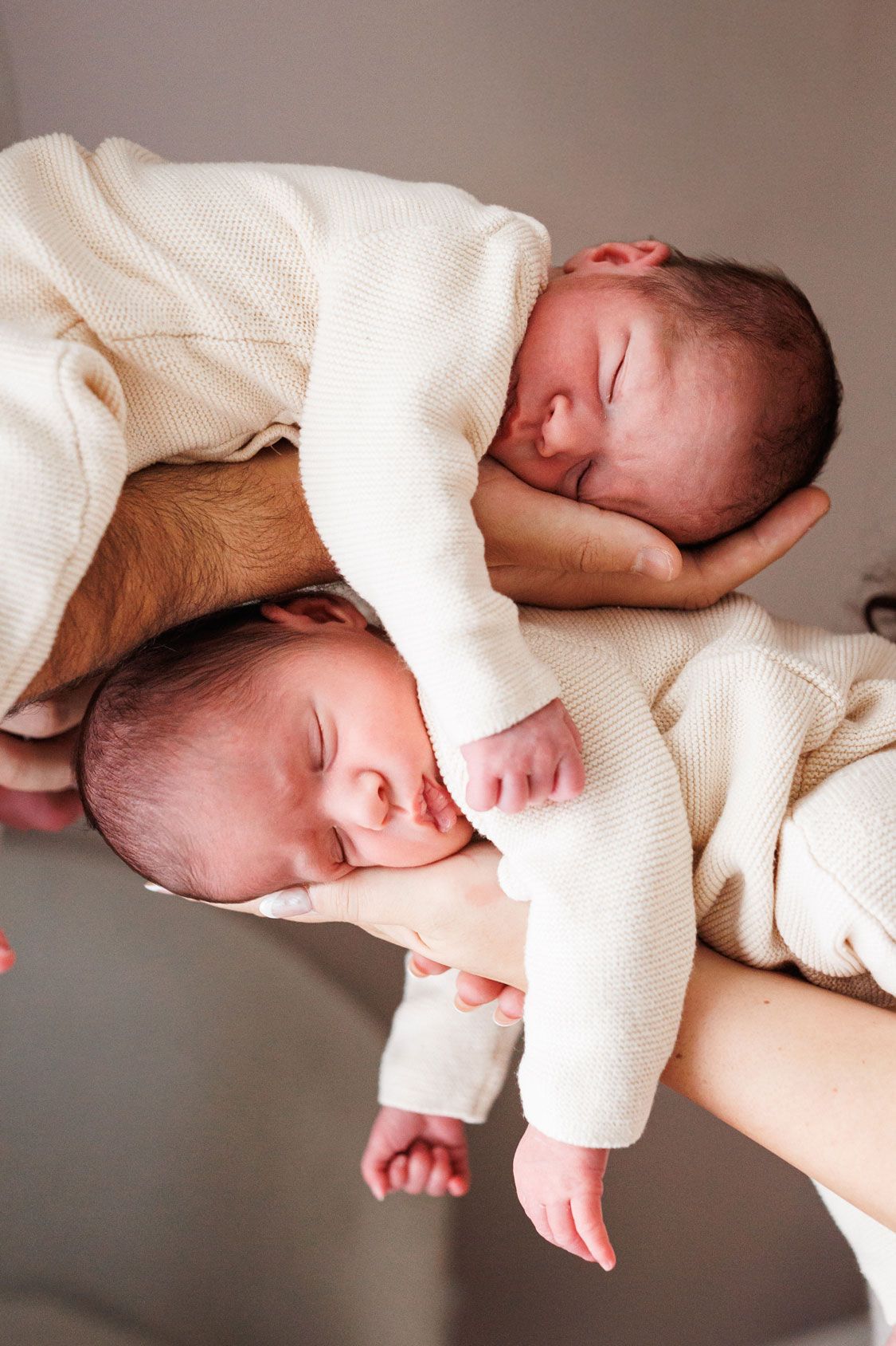 Two sleeping newborn babies in cream knit outfits rest peacefully on the forearms and hands of two parents in a bright, soft-lit room.