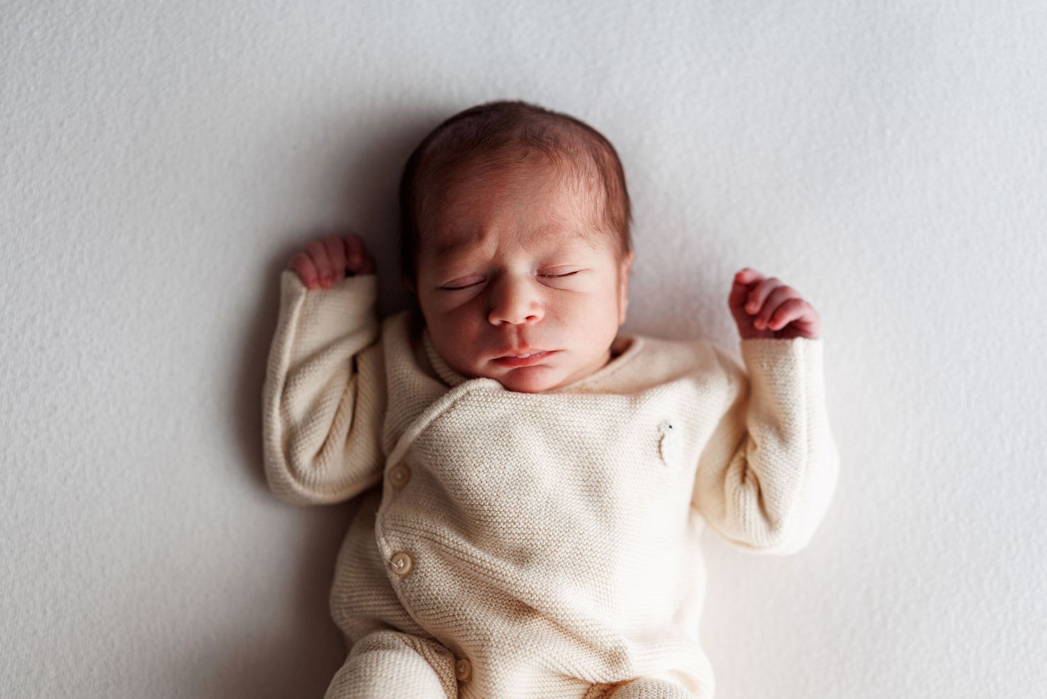 Sleeping newborn baby in a cream wrap-style onesie lying on a soft neutral background with arms raised beside the head.