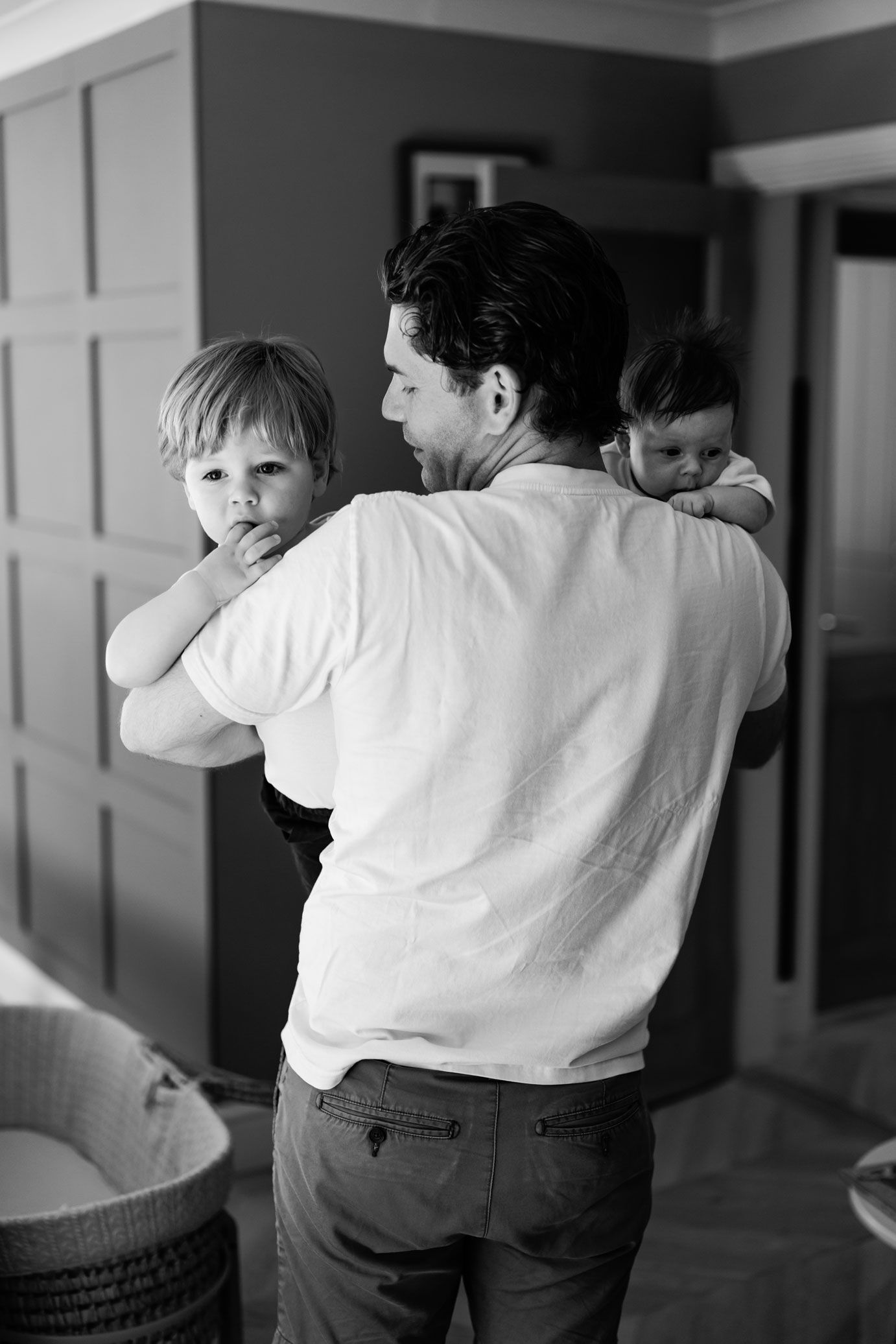 Black and white photo of a father holding two young children, a toddler and baby, in a home interior.