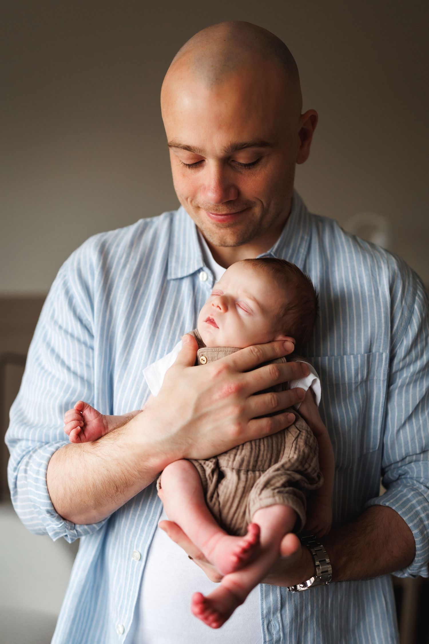 Bald father in blue shirt smiling warmly while cradling his newborn baby against his chest.
