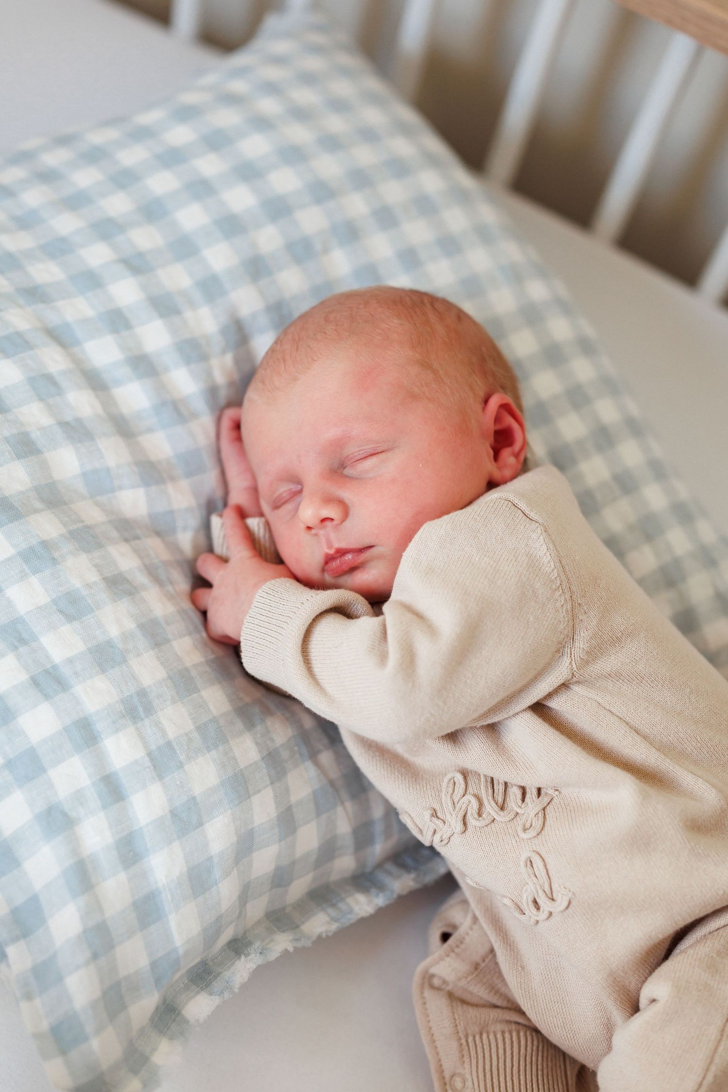 Sleeping newborn baby in a cream knit outfit resting peacefully on a blue and white gingham pillow in a white crib.