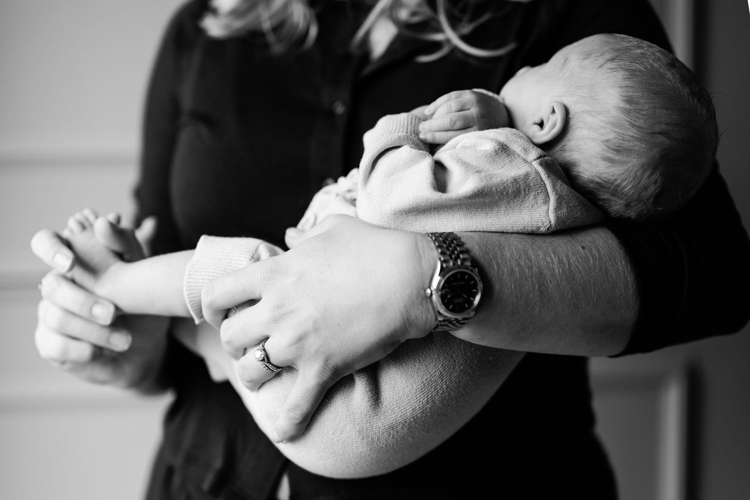 Black and white closeup of a woman wearing a wristwatch and ring tenderly holding a swaddled newborn baby in her arms.