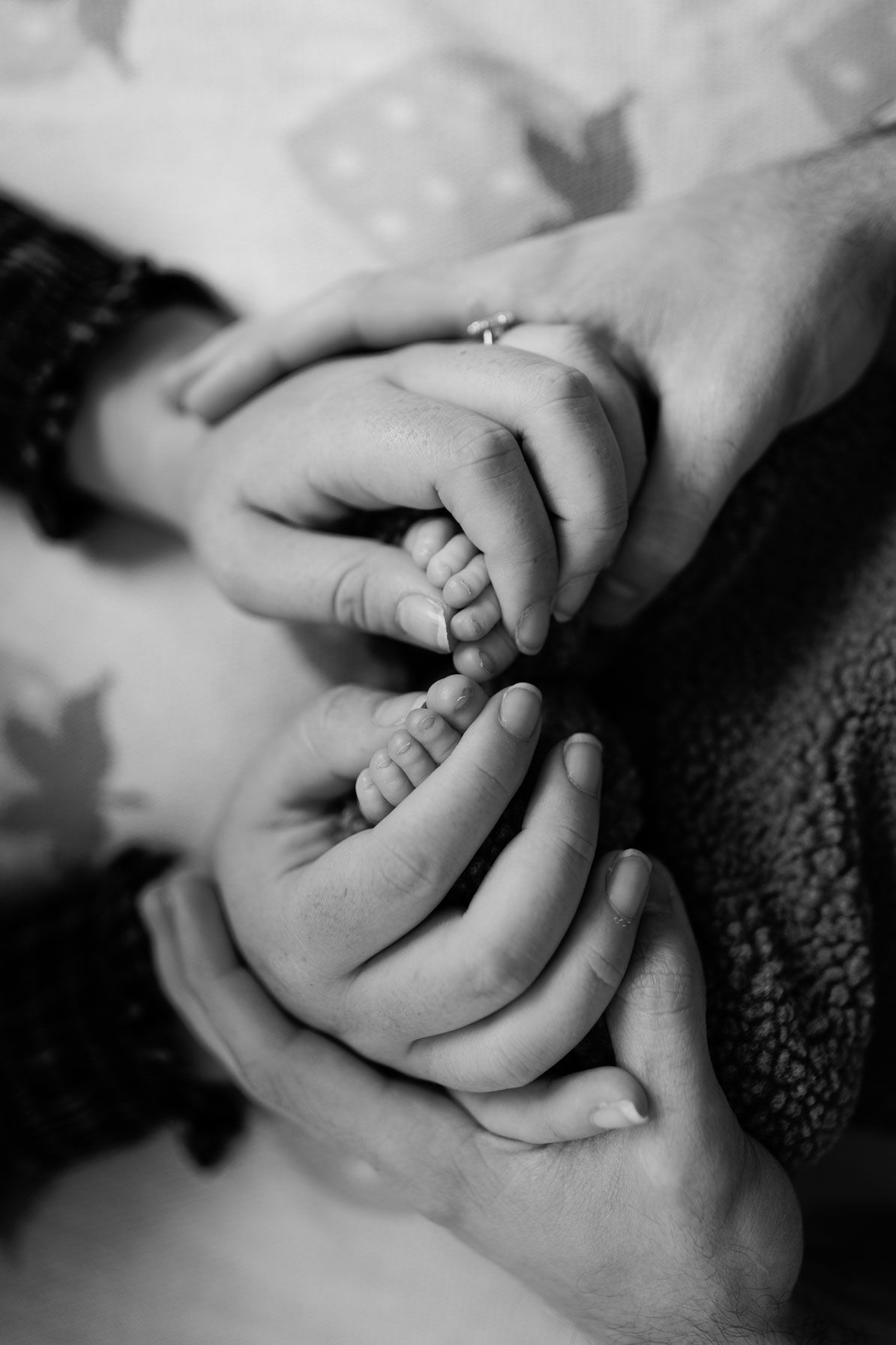 Black and white photo of two adult hands gently cradling a newborn baby's tiny feet, one hand wearing a ring.