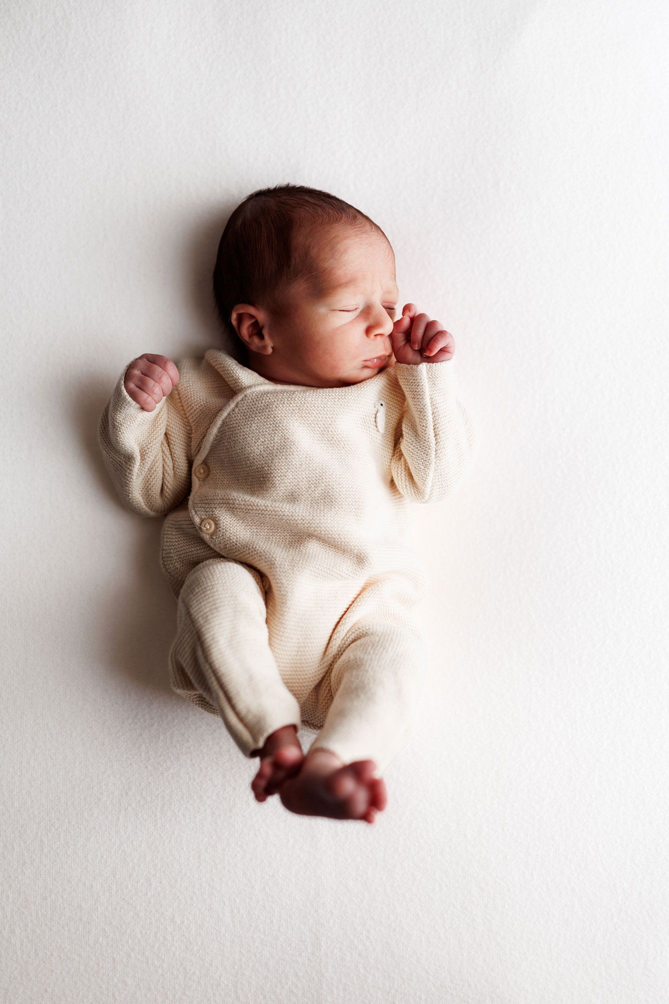 Sleeping newborn baby in a cream knit button outfit lying on a soft white background with tiny fists raised near the face.