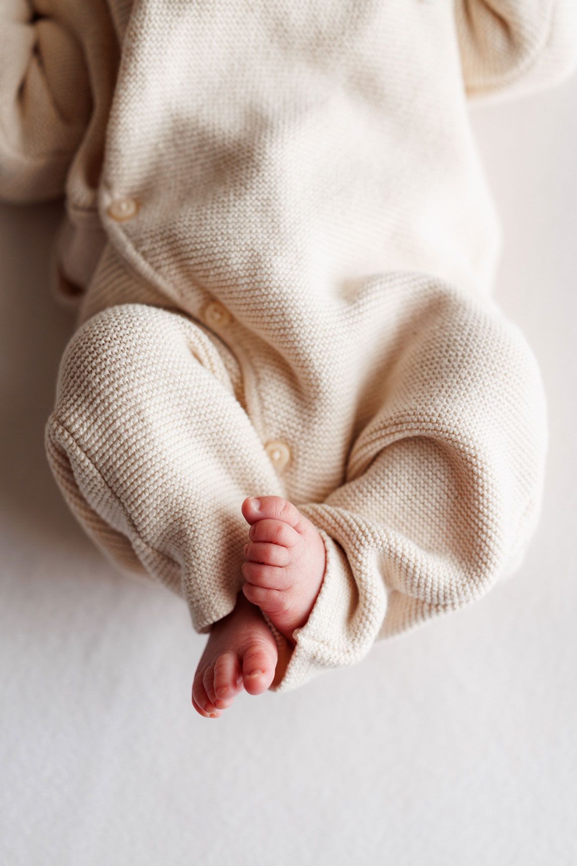 Tiny newborn baby feet peeking out from a cream knit button-up romper against a soft white background.