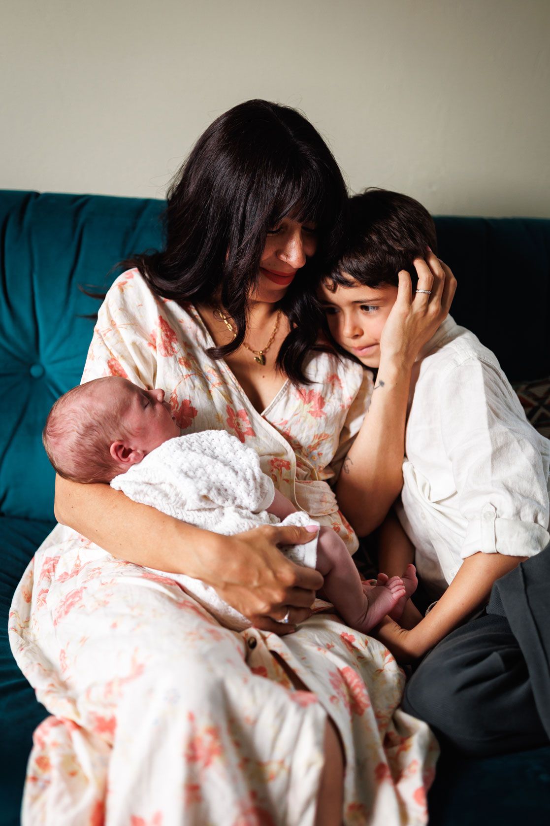 Mother in floral dress sitting on teal sofa cuddling newborn and young child.