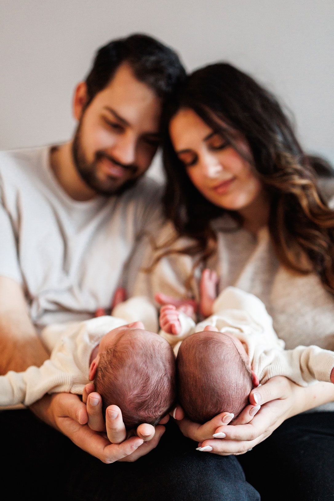 Smiling parents sit close together gazing down at their two sleeping newborn babies held side by side in their hands.