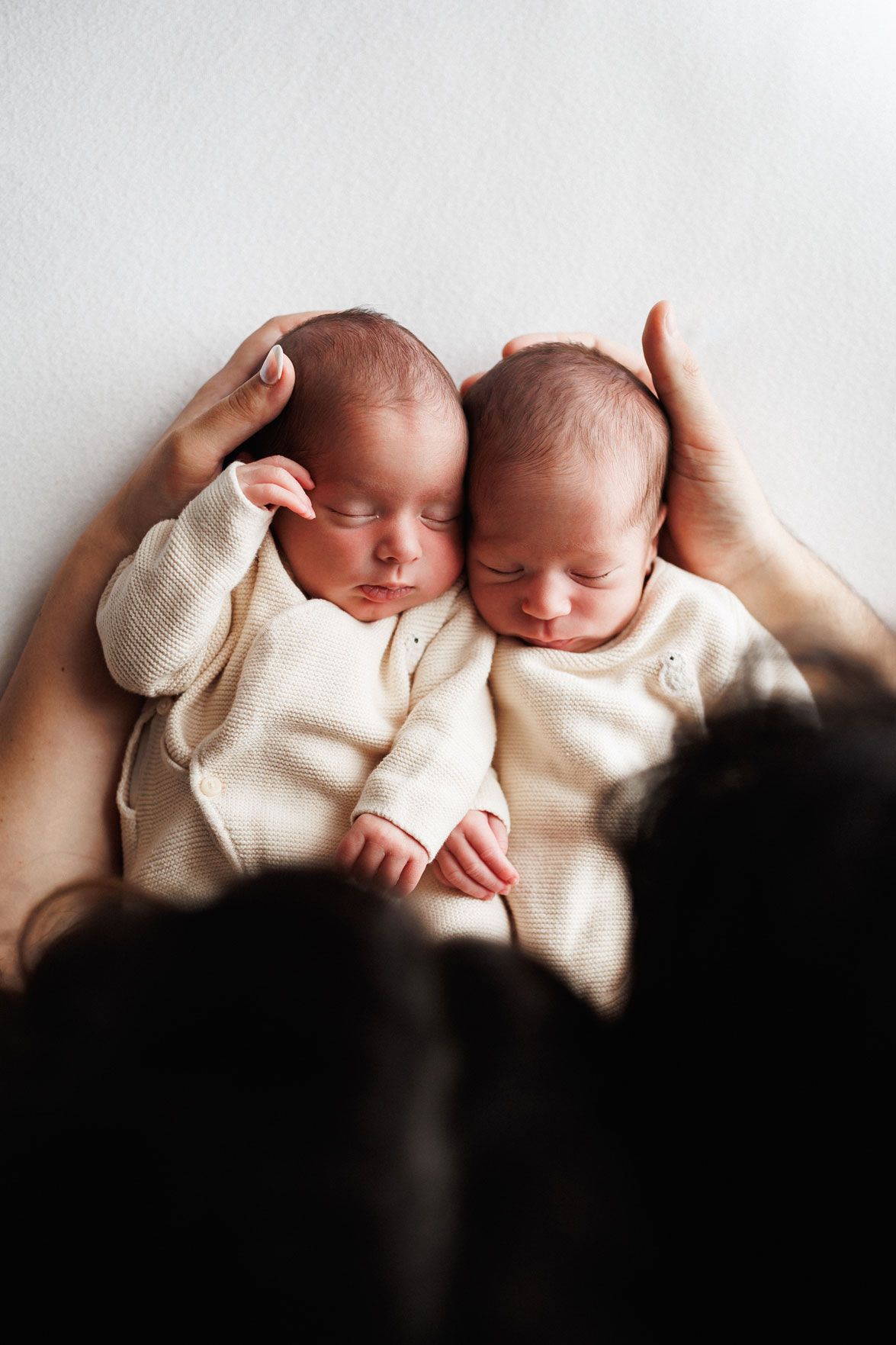Two sleeping newborn twin babies in cream outfits cuddled together while a parent's hands gently cradle their heads.