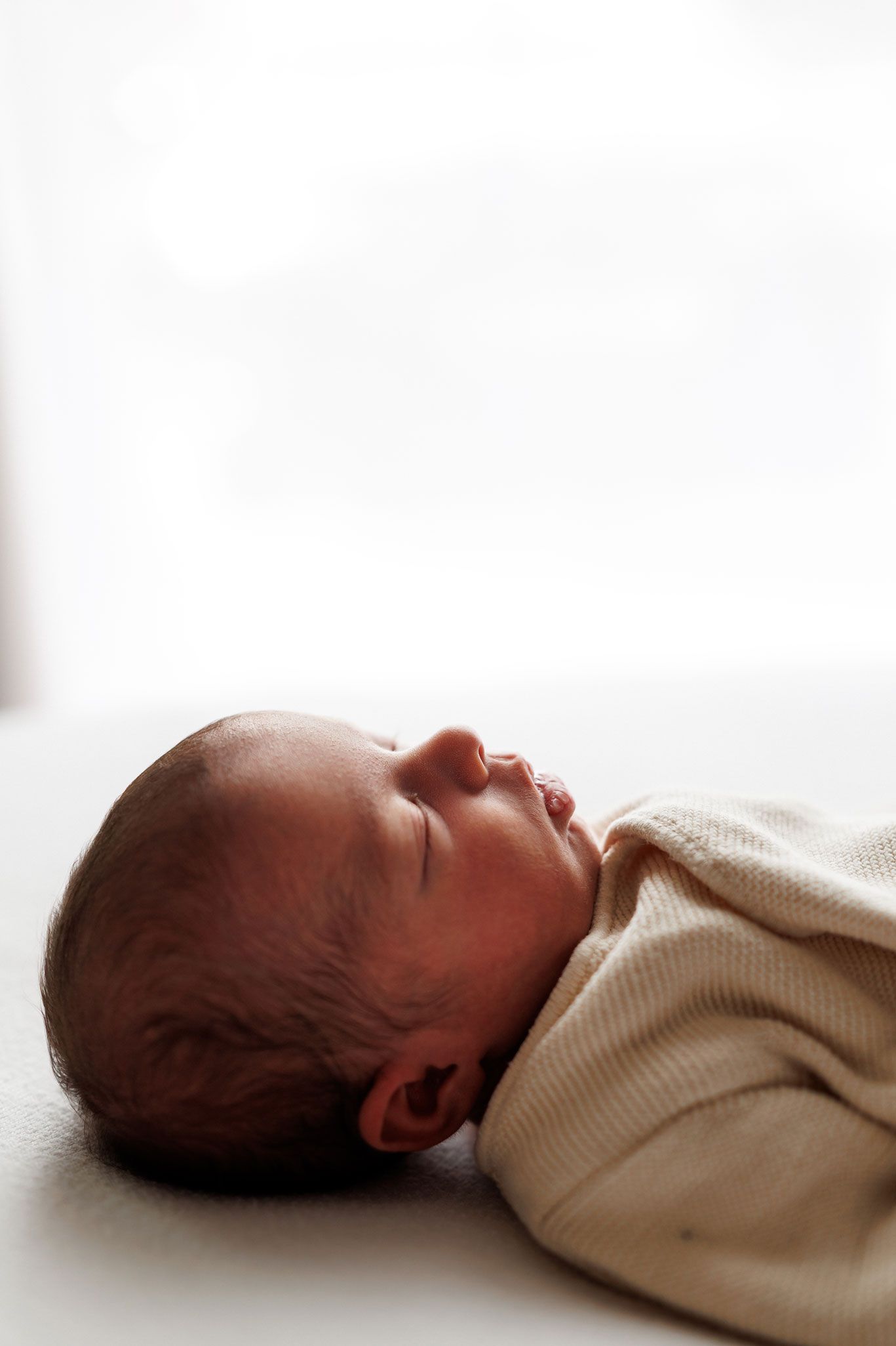 Side profile of a sleeping newborn baby in a cream knit outfit resting peacefully against a soft, bright white background.