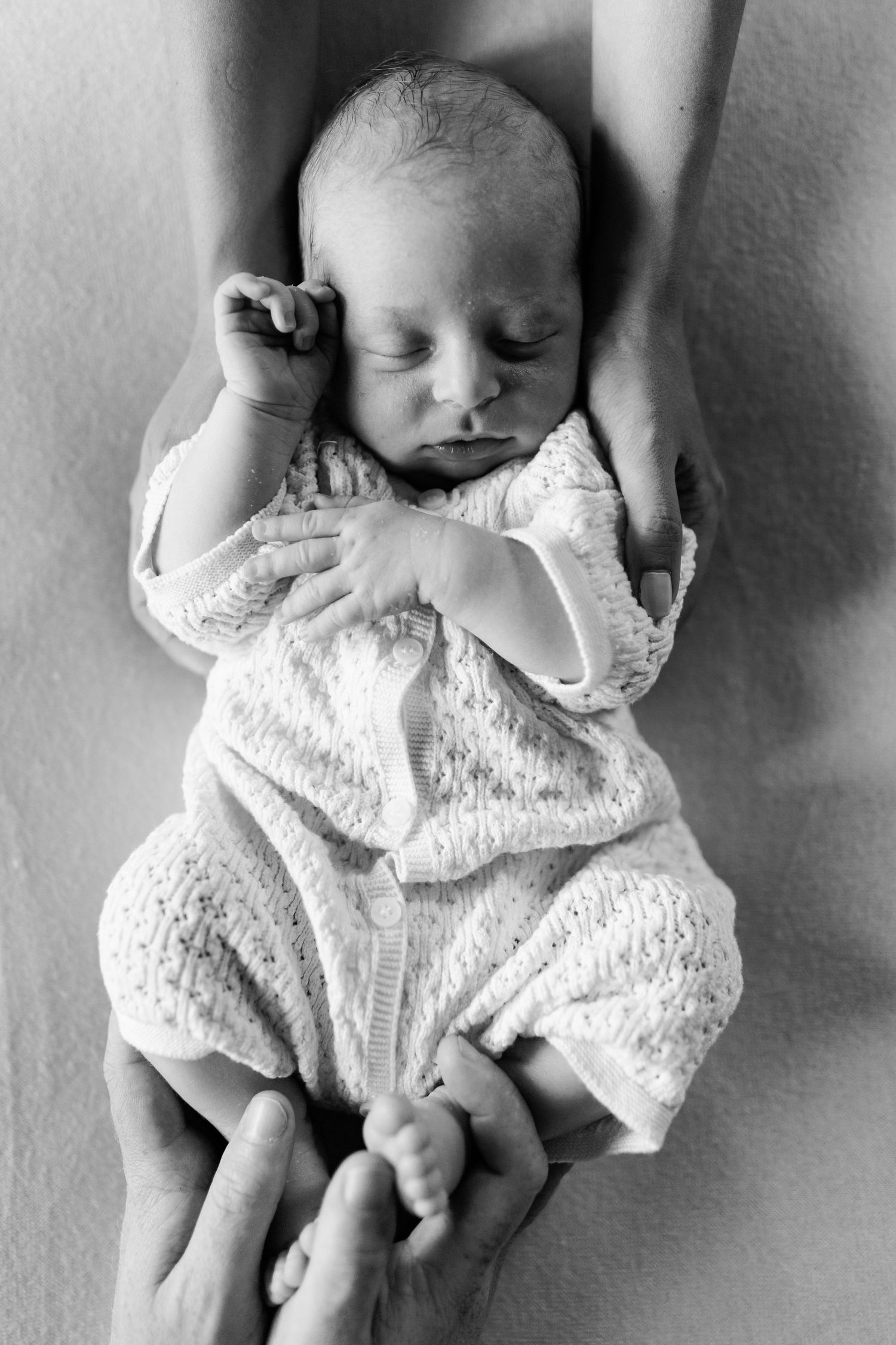 Black and white overhead view of sleeping newborn baby in white knit outfit held by parents.