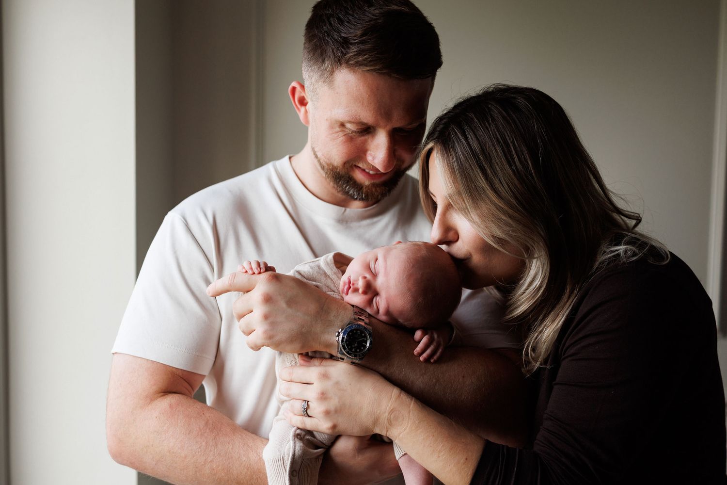 Smiling father in a white t-shirt and mother kissing their sleeping newborn baby's head in a bright, cozy home newborn photo.