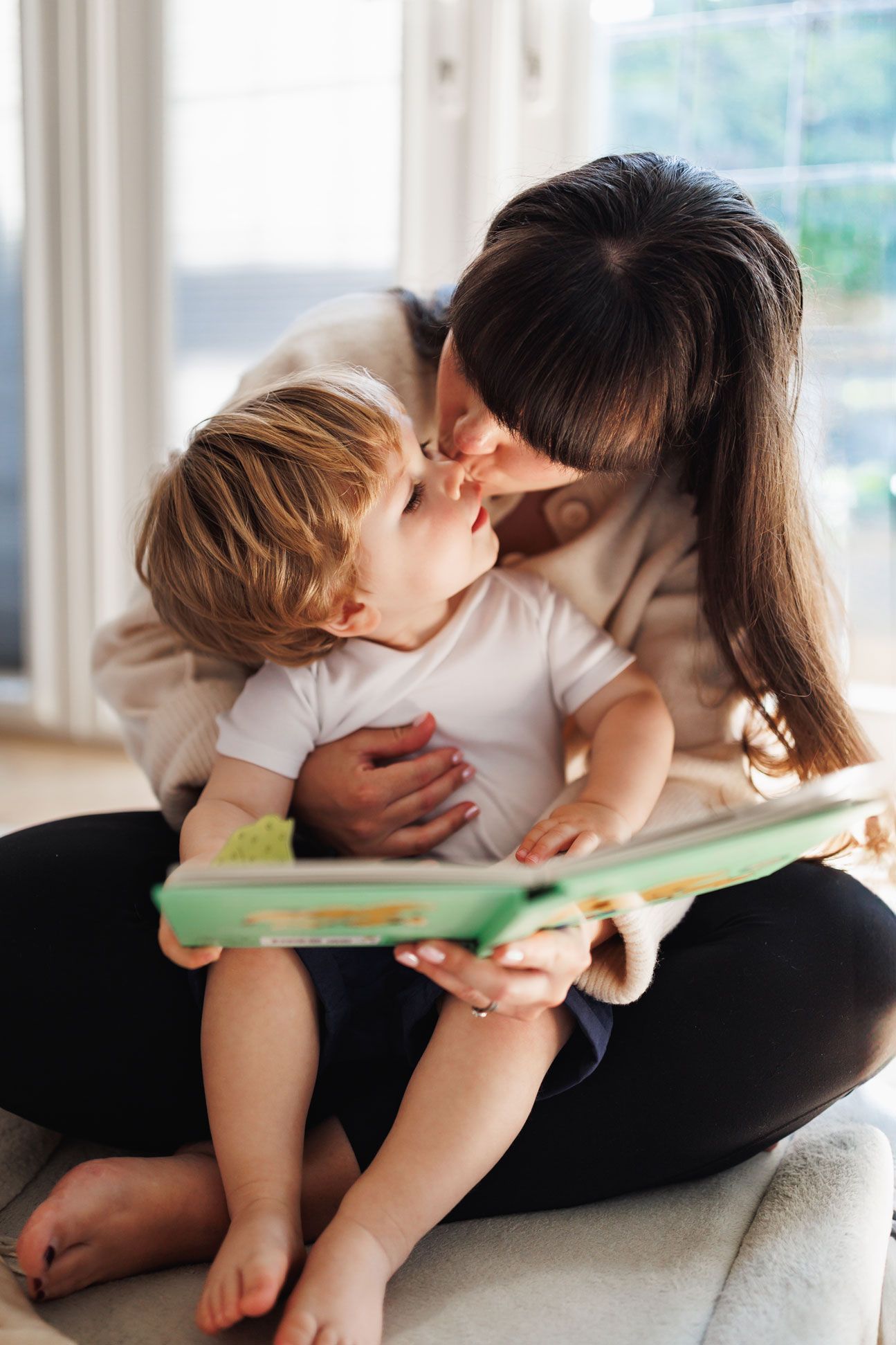 A mother sits cross-legged reading a picture book while sharing a tender nose kiss with her toddler.
