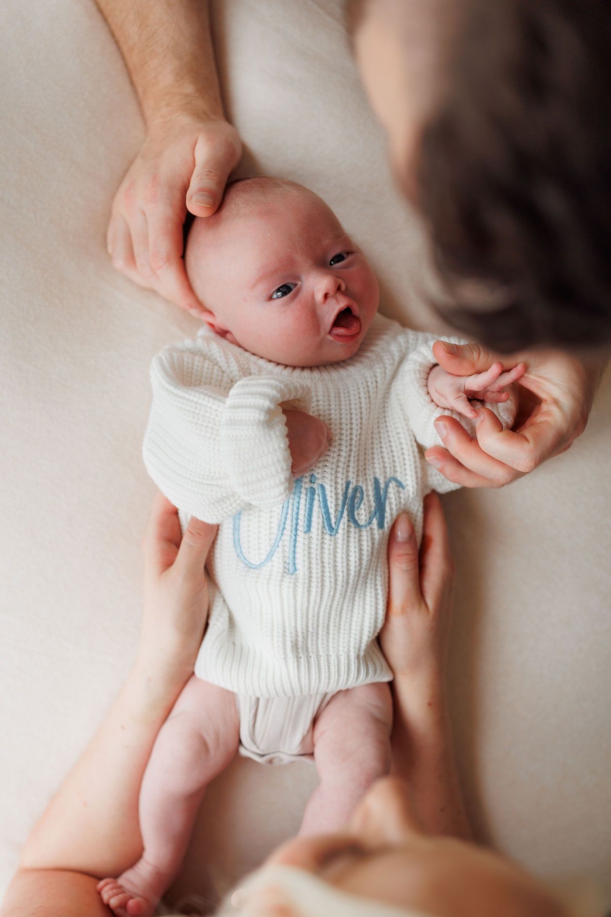 Newborn baby named Oliver in white knit onesie being held by parents on a soft white surface.