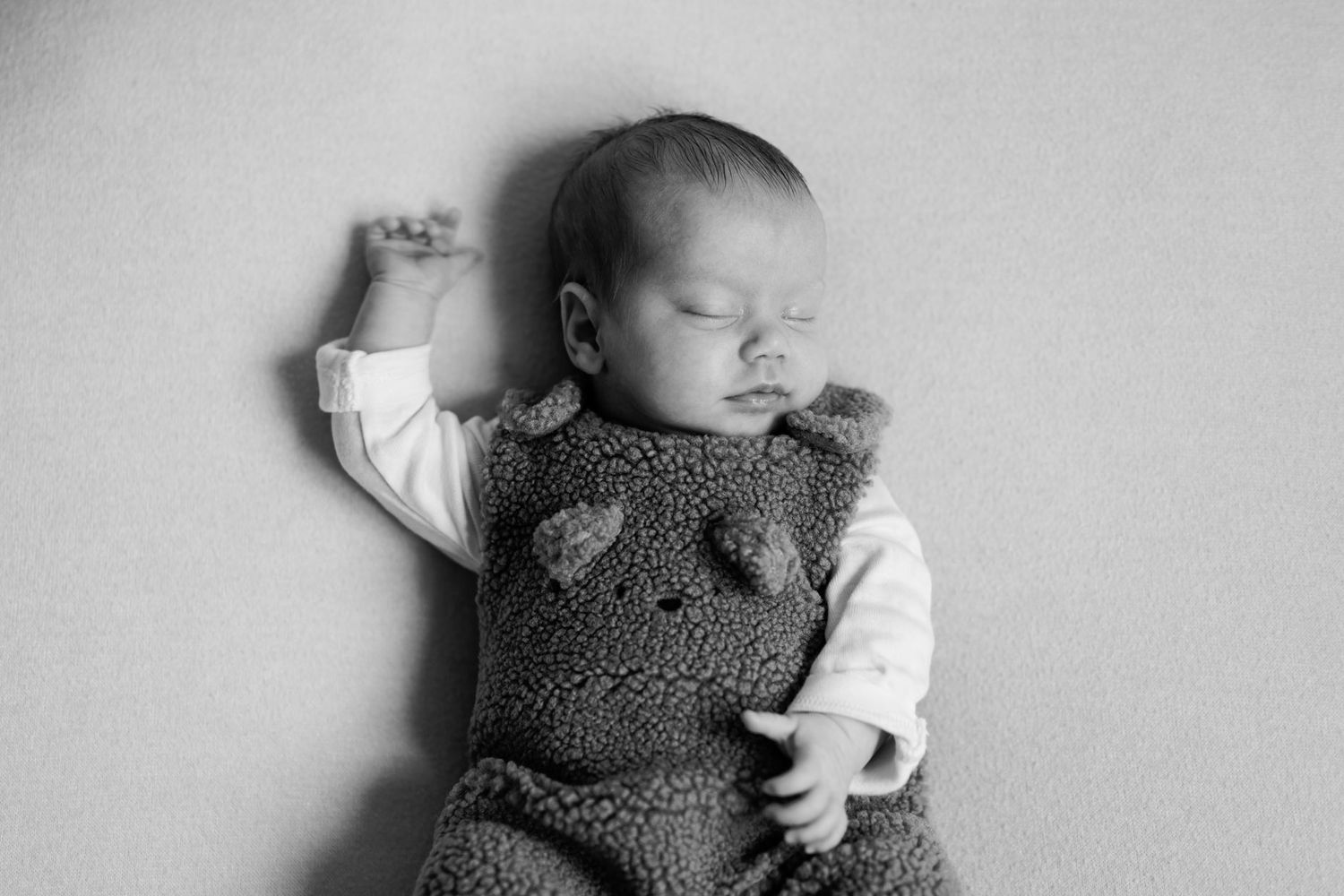 Black and white photo of a sleeping newborn baby in a fuzzy bear-ear romper with one arm raised, resting peacefully on a soft surface.
