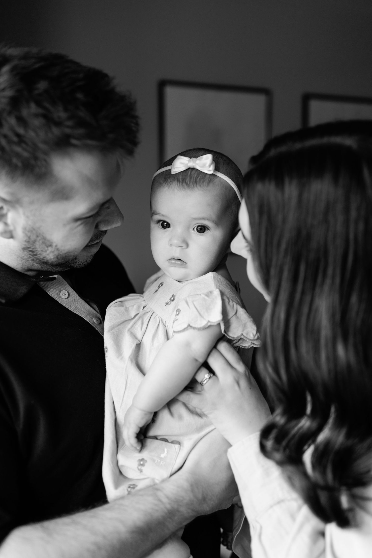 Black and white family portrait of a man and woman gazing lovingly at a baby girl wearing a bow headband and a smocked dress.