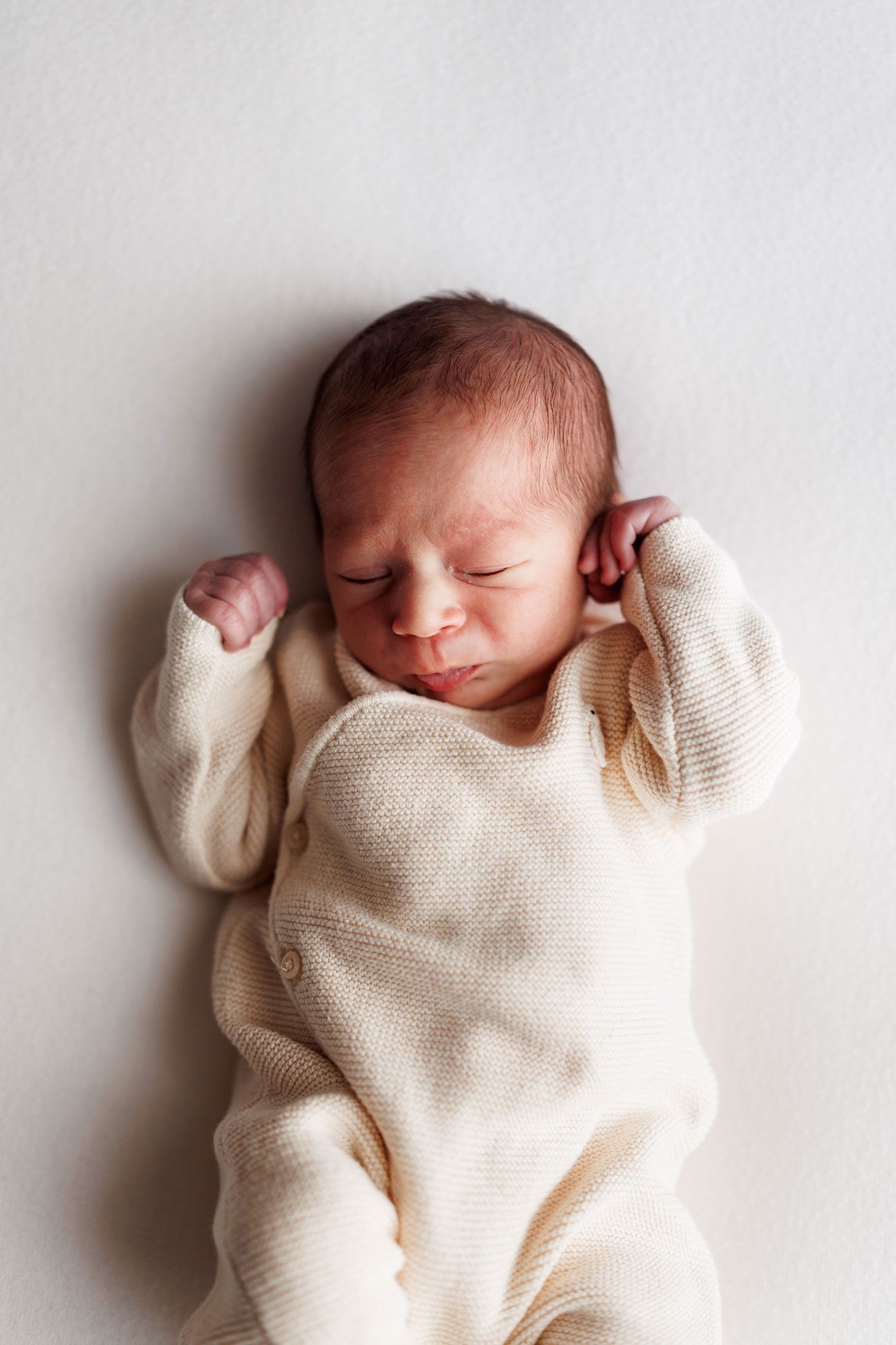 Sleeping newborn baby in a cream knit outfit lying on a soft white background with tiny hands raised near the face.