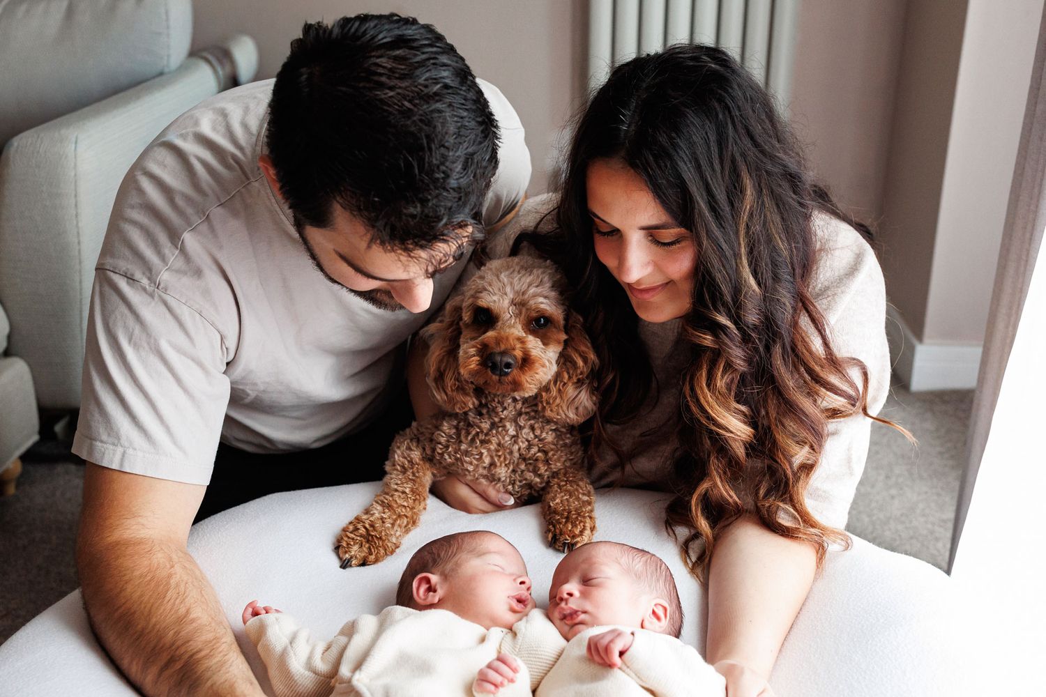 A man and woman smile down at two newborn twin babies lying side by side while a fluffy brown goldendoodle dog sits between them.