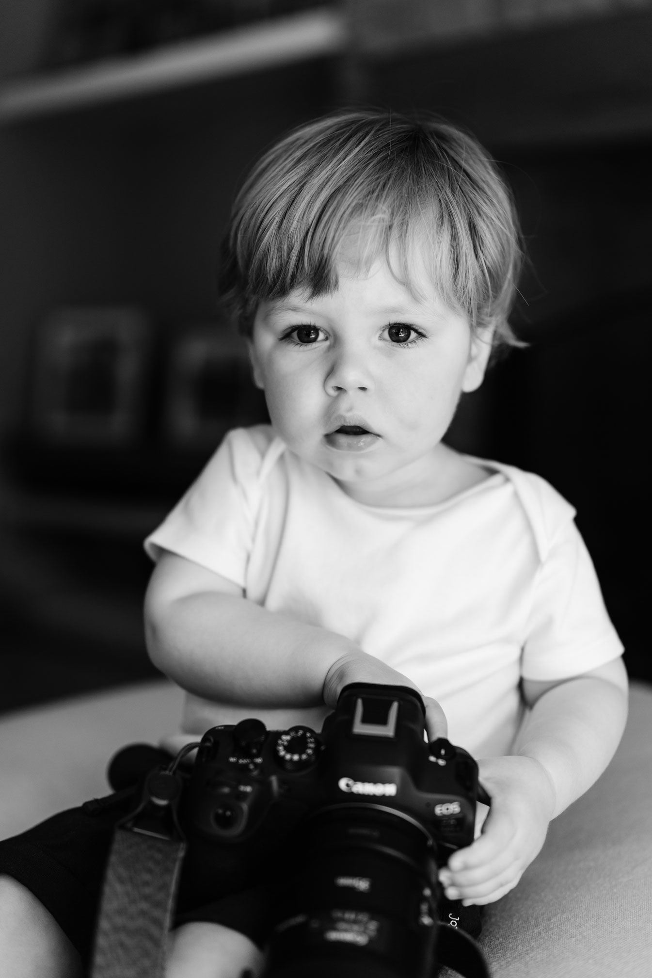 Curious blonde toddler in white onesie holds a Canon DSLR camera in black and white portrait photo.