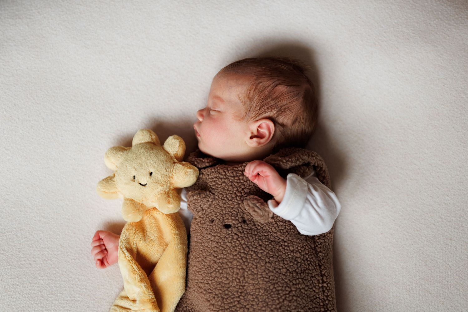 Sleeping newborn baby in a brown fleece sleeper lying on a cream blanket beside a soft yellow sun-shaped plush comforter toy.