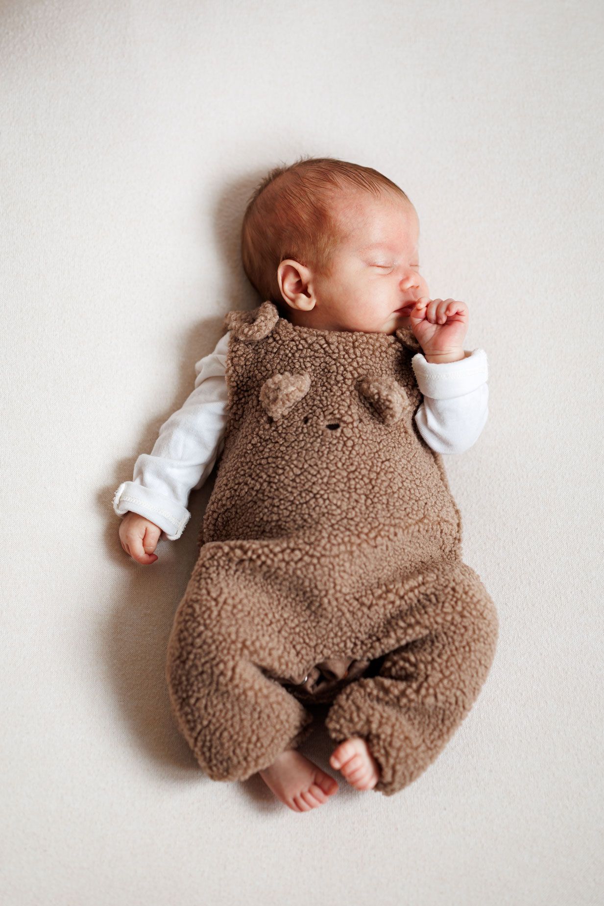 Newborn baby sleeping peacefully on a cream blanket, wearing a brown teddy bear fleece romper with ears and a white long-sleeve bodysuit.
