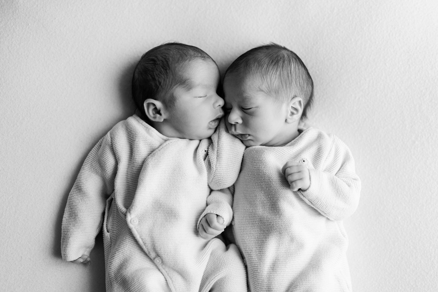 Black and white photo of two sleeping newborn twins in matching white outfits lying side by side with their heads touching.