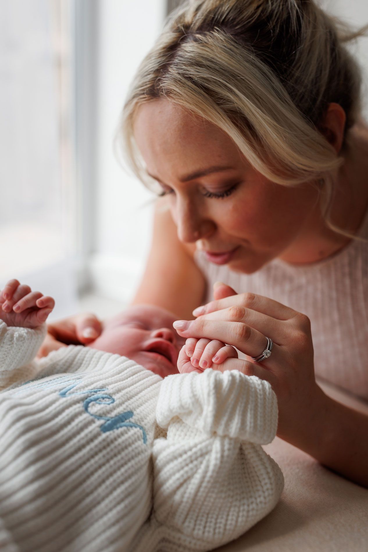A tender moment as a blonde mother gazes lovingly at her newborn baby dressed in a white knit outfit near a window.