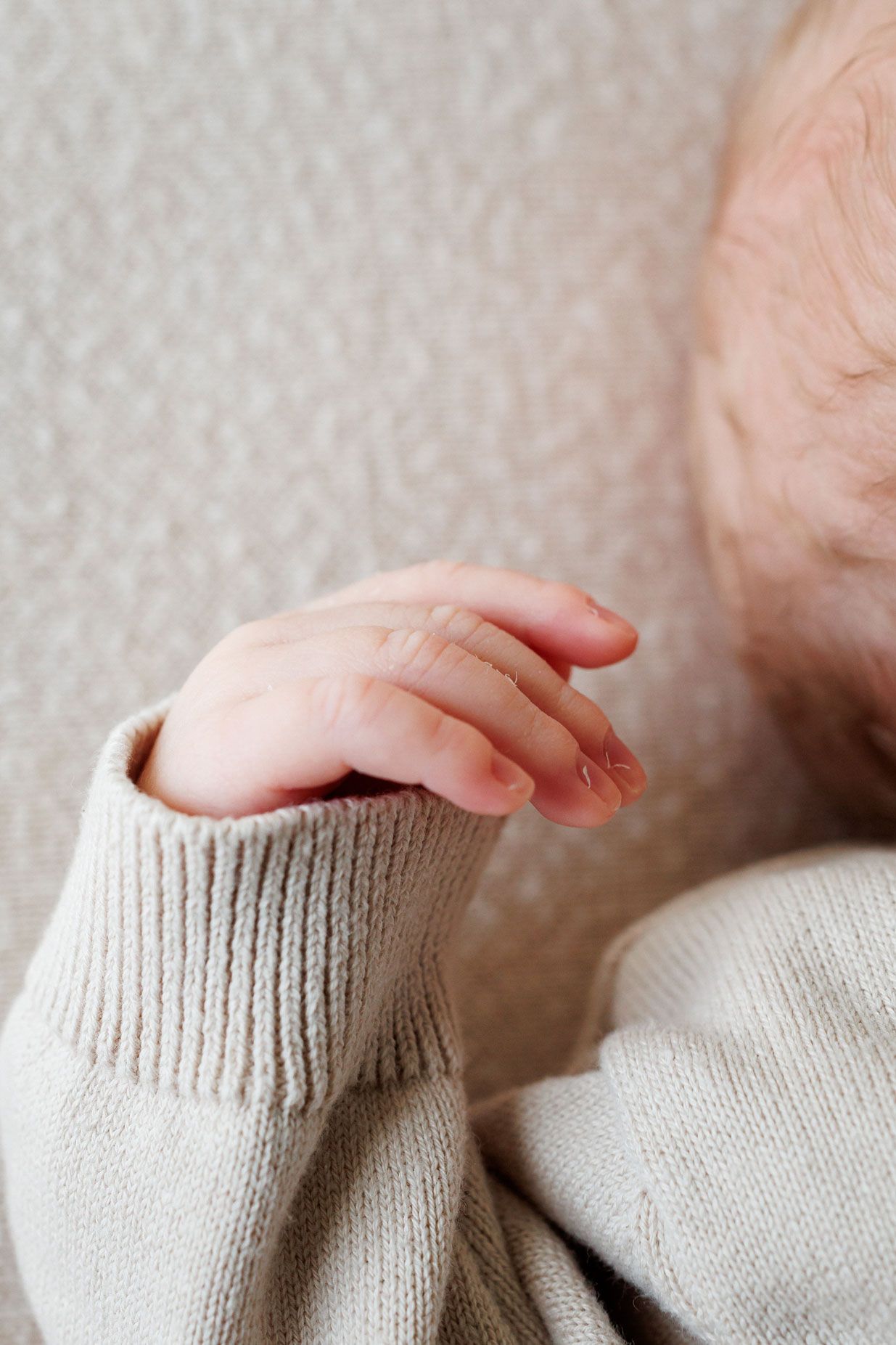 Close-up of a sleeping newborn's tiny hand with delicate fingers resting on a soft beige blanket, wearing a cozy cream knit sweater.